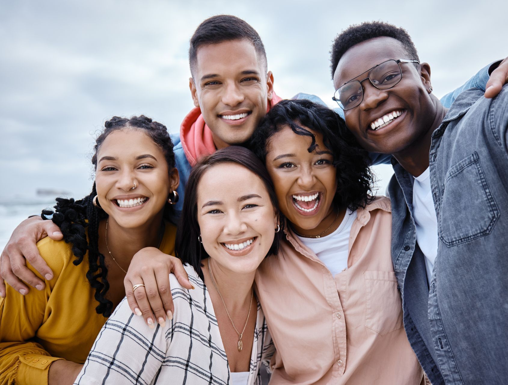 A diverse group of five smiling friends huddle closely together for a bright, casual outdoor selfie.