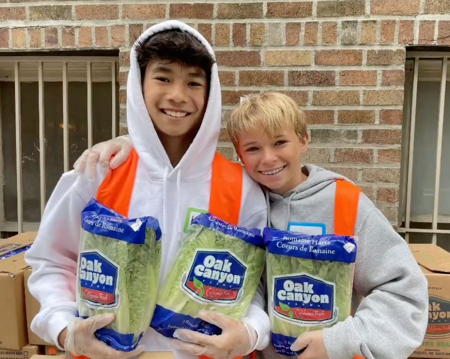 Two kids in orange safety vests holding up bags of lettuce during a volunteer service opportunity. One is wearing a hoodie and has their hood on.