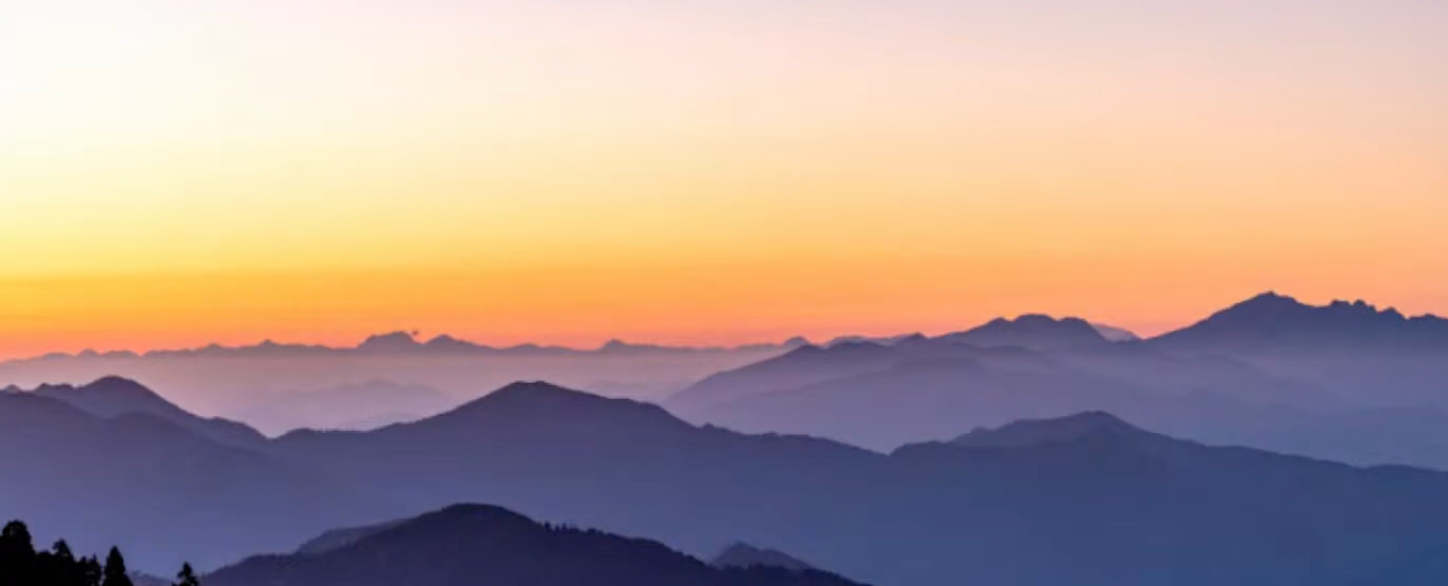 A vast purple mountain range covered in fog at dusk. The sun is setting a bright gold sunset behind.