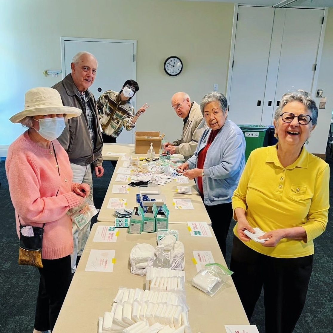 Senior adults helping pack hygiene kits for the Hope Center. Many different hygiene-related items on the table. People are smiling at the camera