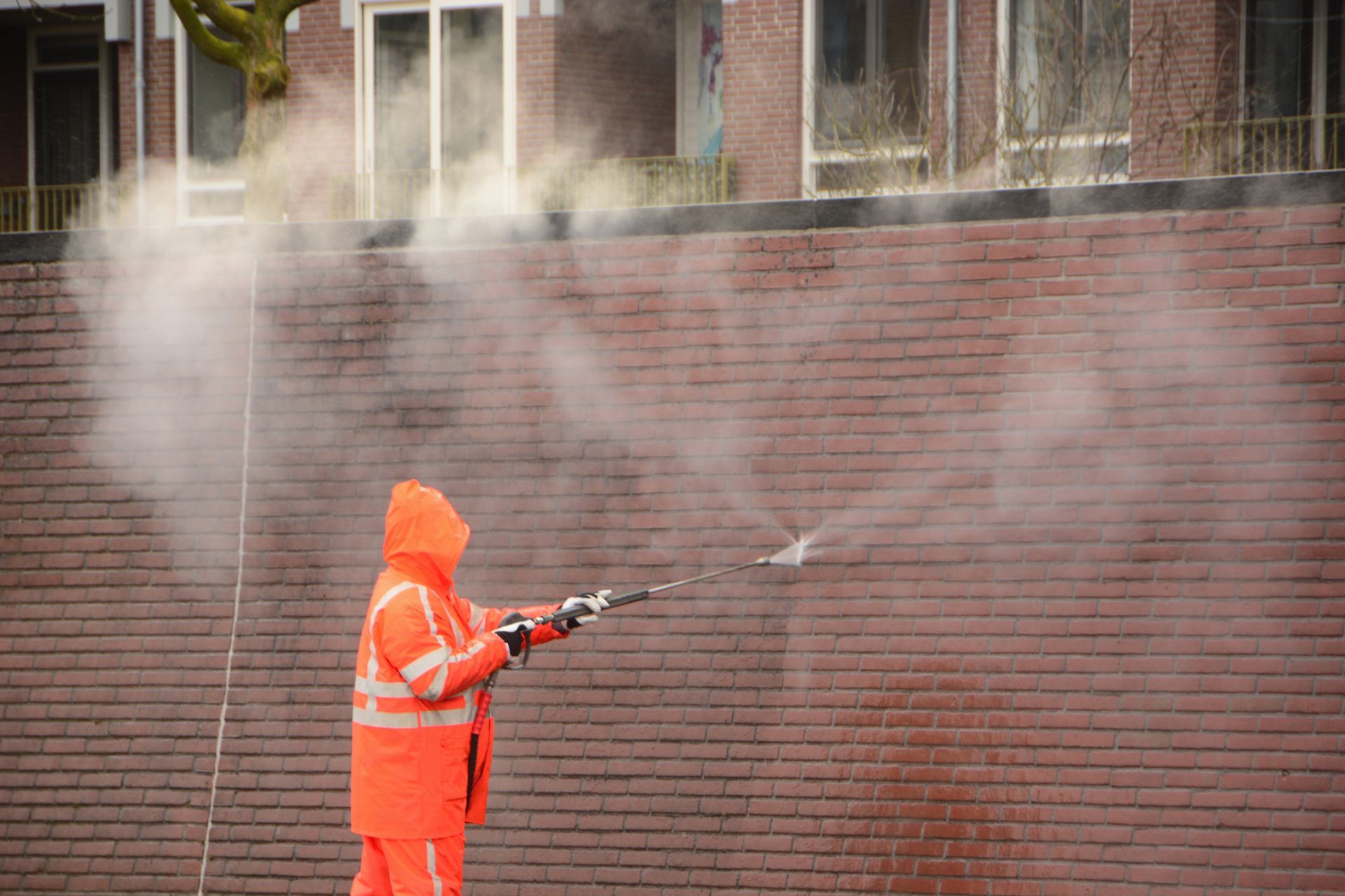 Worker in orange suit pressure washing a brick wall with steam.