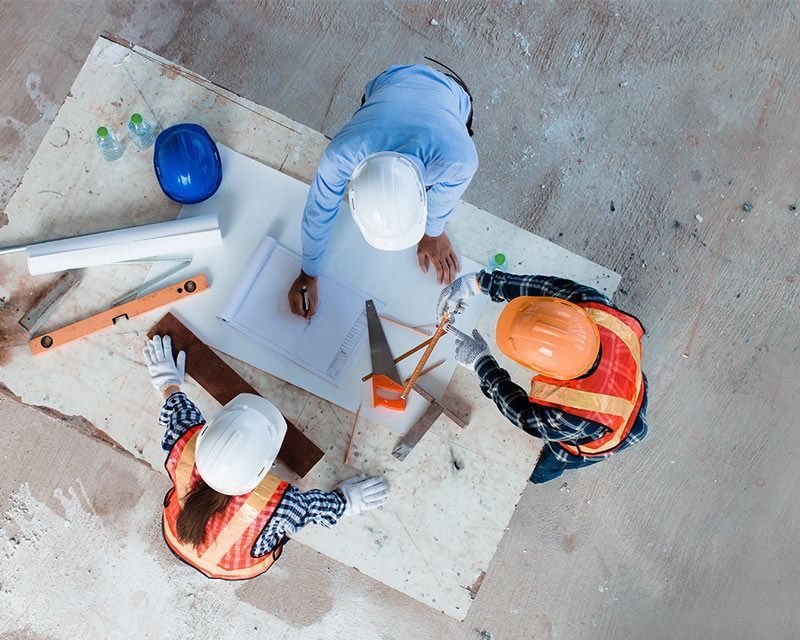 Construction workers in hard hats reviewing plans around a blueprint on a floor.