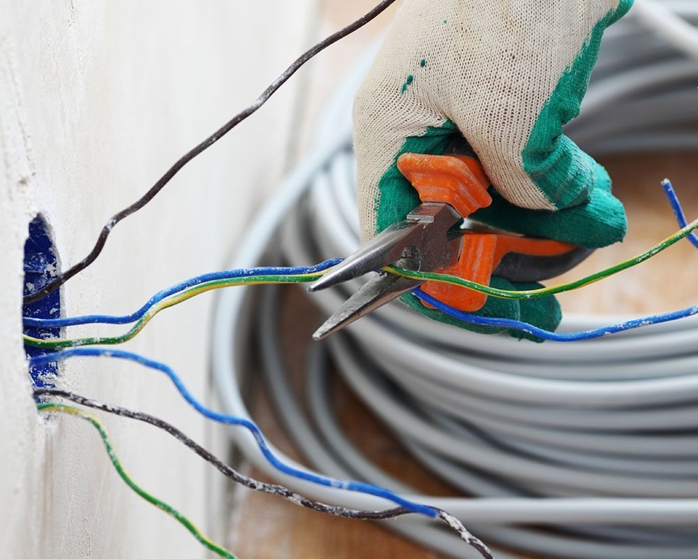 Gloved hand stripping blue and green electrical wires with pliers near a wall outlet area