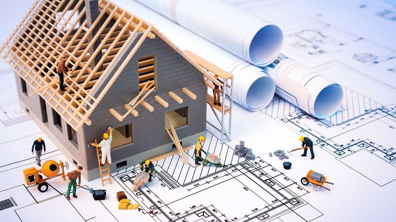 Miniature house under construction on blueprints, with workers and tools around it