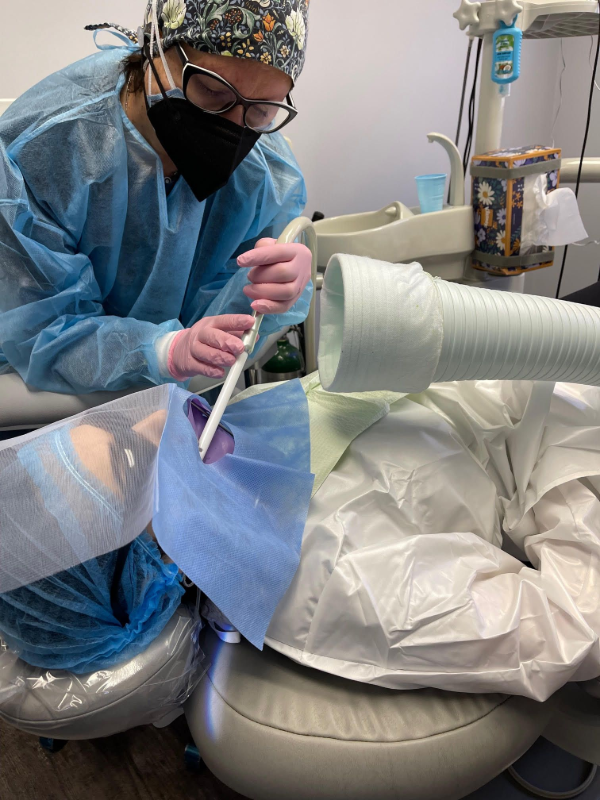 Dentist in blue protective gear examines a patient reclined in a dental chair
