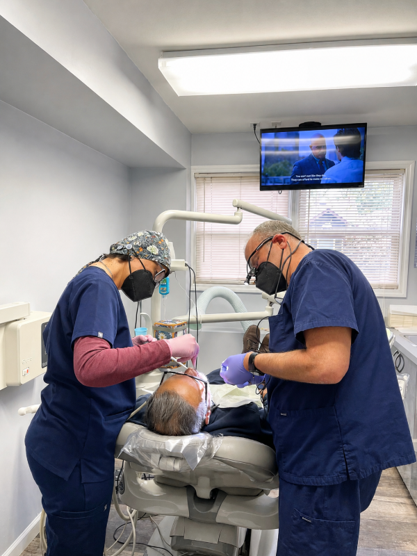 Two dentists treating a patient in a bright dental exam room, with a TV mounted above the chair.