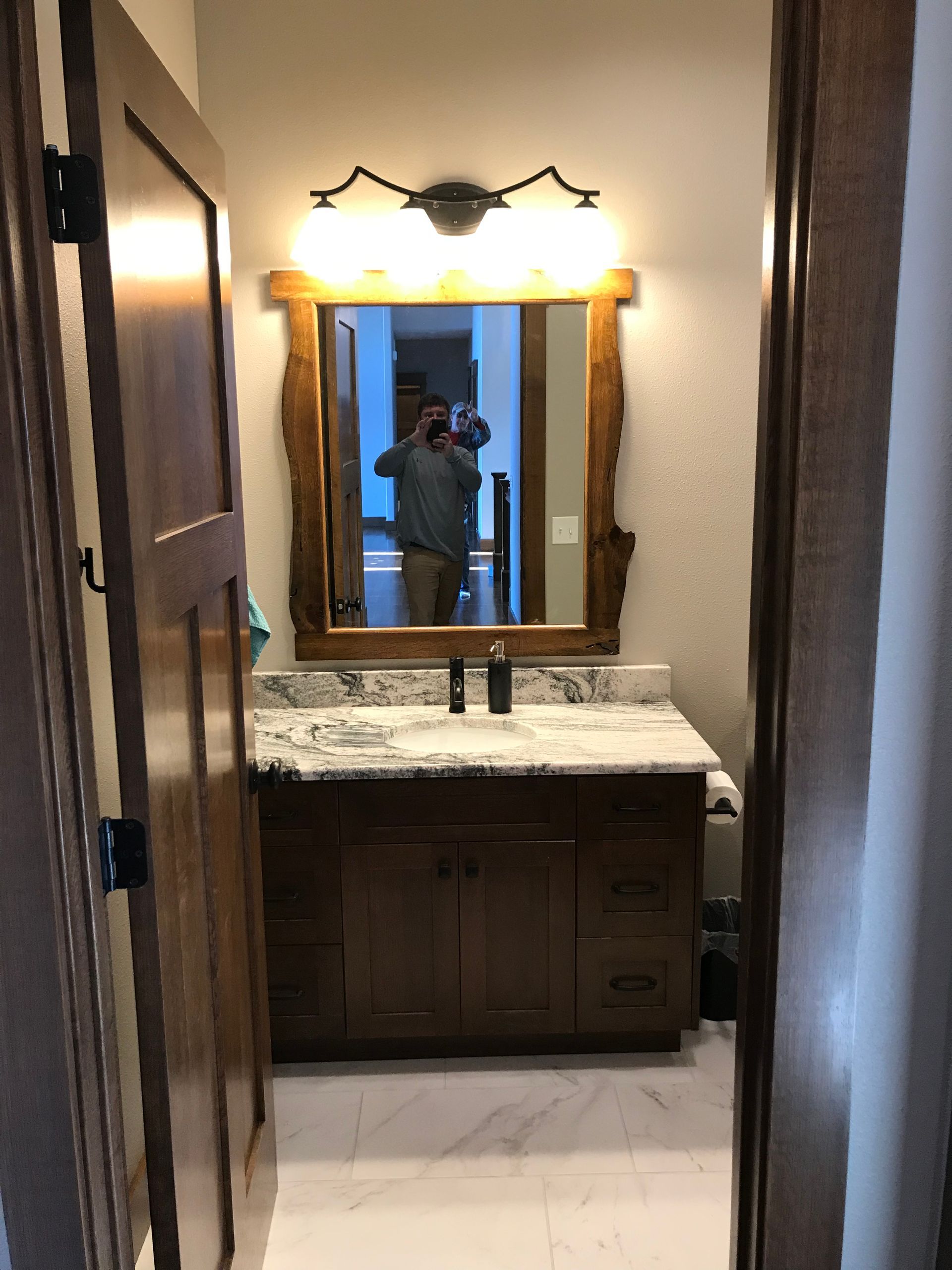 Narrow bathroom with a granite vanity, framed mirror, and bright light reflecting a person taking the photo.