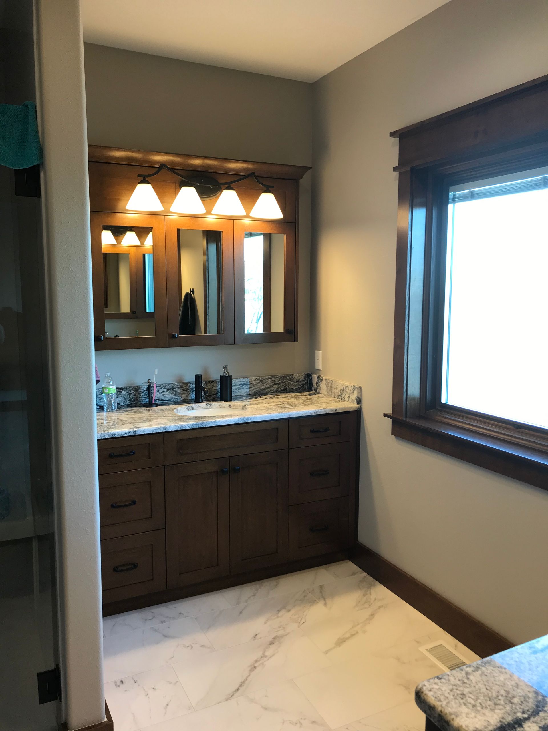 Bathroom vanity with dark wood cabinets, granite countertop, lit mirror, and window with dark trim