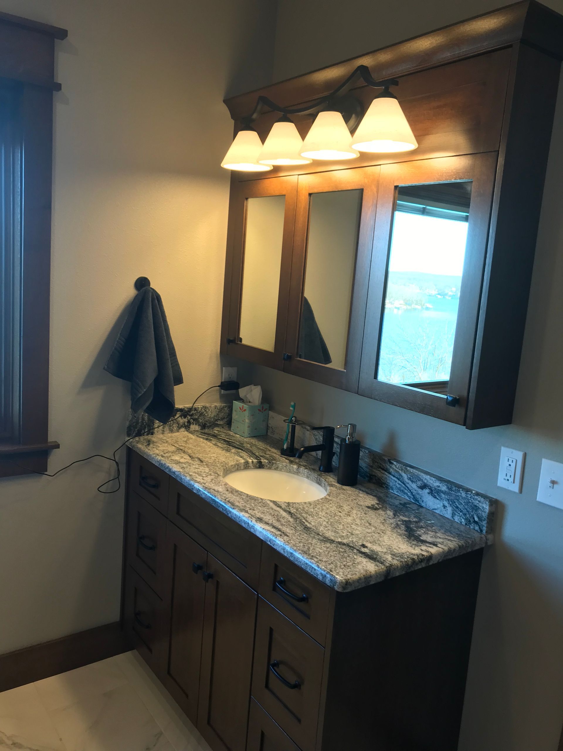 Bathroom vanity with granite countertop, lit triple-light mirror, and towel on the wall