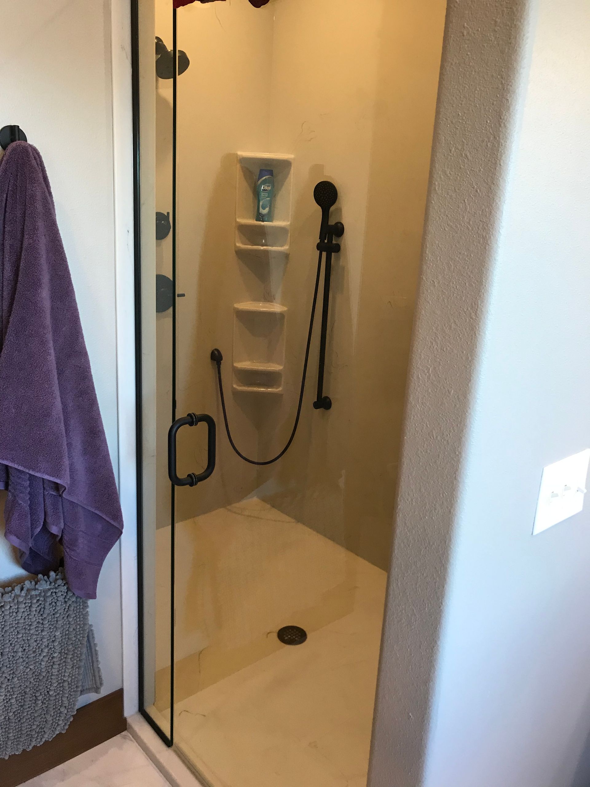 Small beige shower stall with glass door, built-in shelves, and a handheld showerhead with hose.