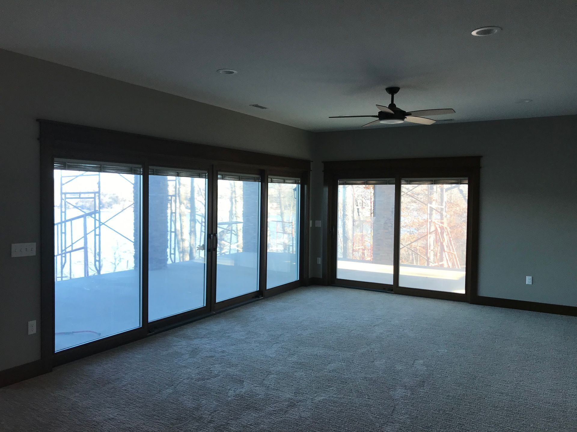 Empty living room with large windows, gray carpet, and a ceiling fan
