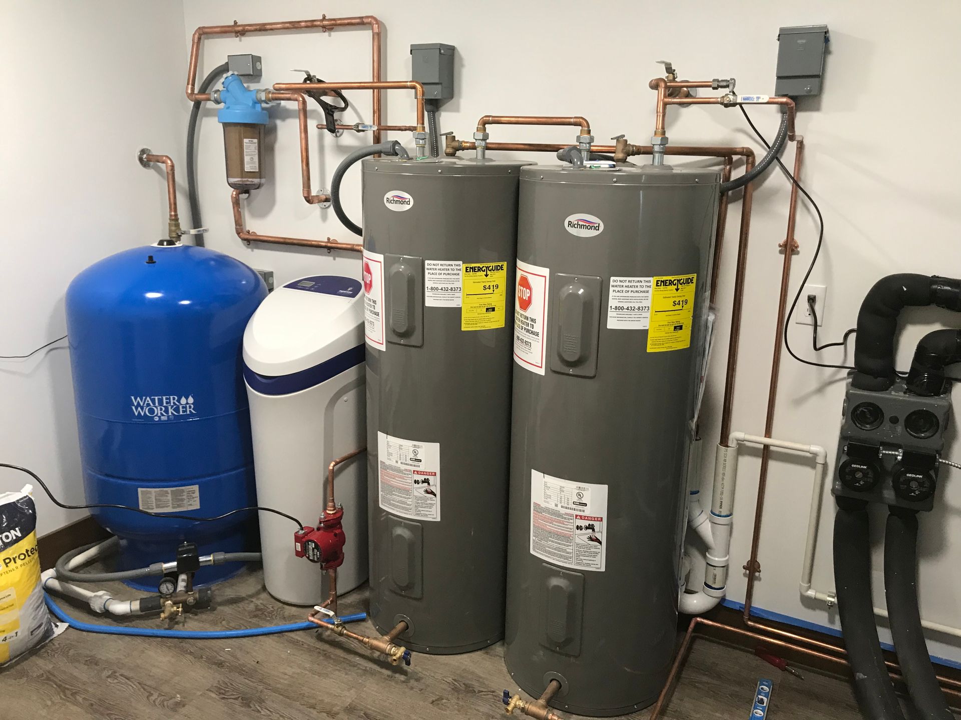 Utility room with two gray water heaters, a blue pressure tank, and copper pipes against a wall