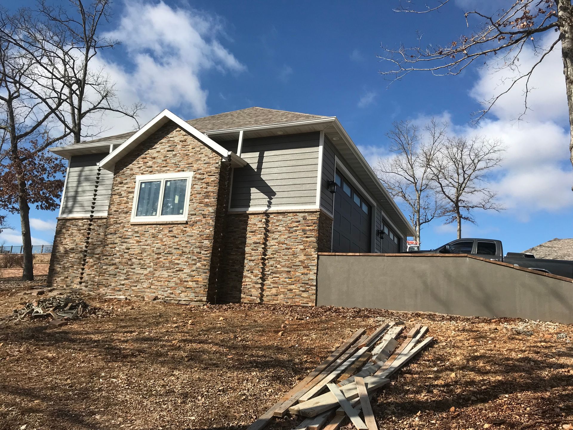 Single-story house with stone and siding, leafless trees, and a wooden ramp in a dry yard under a blue sky