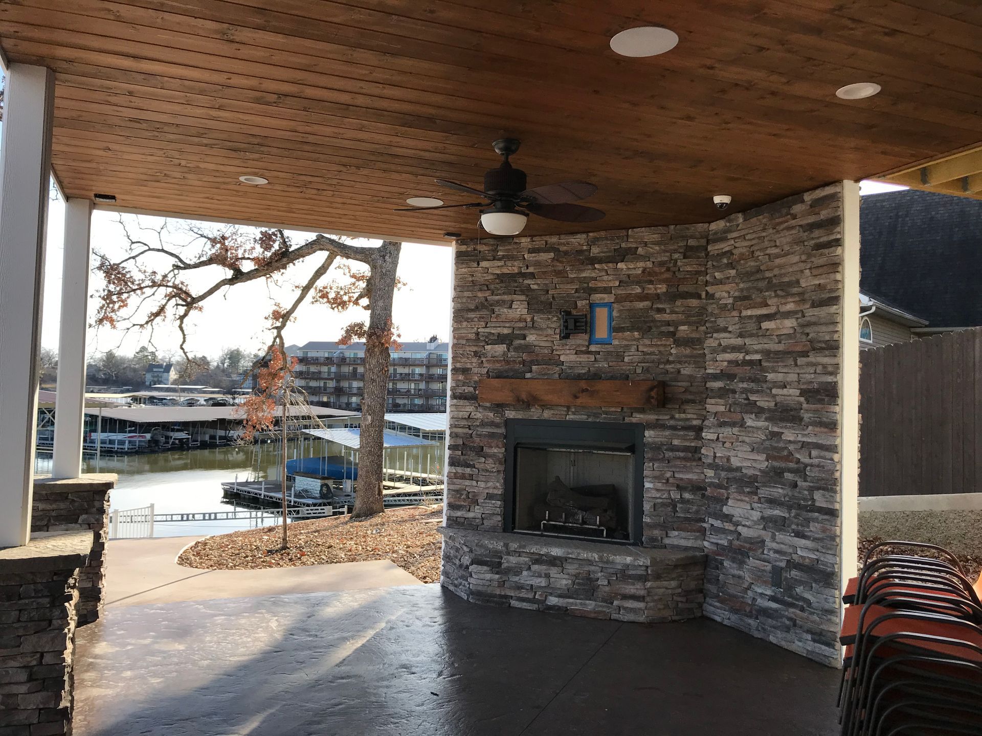 Covered outdoor patio with stone fireplace and wooden ceiling overlooking a waterfront.