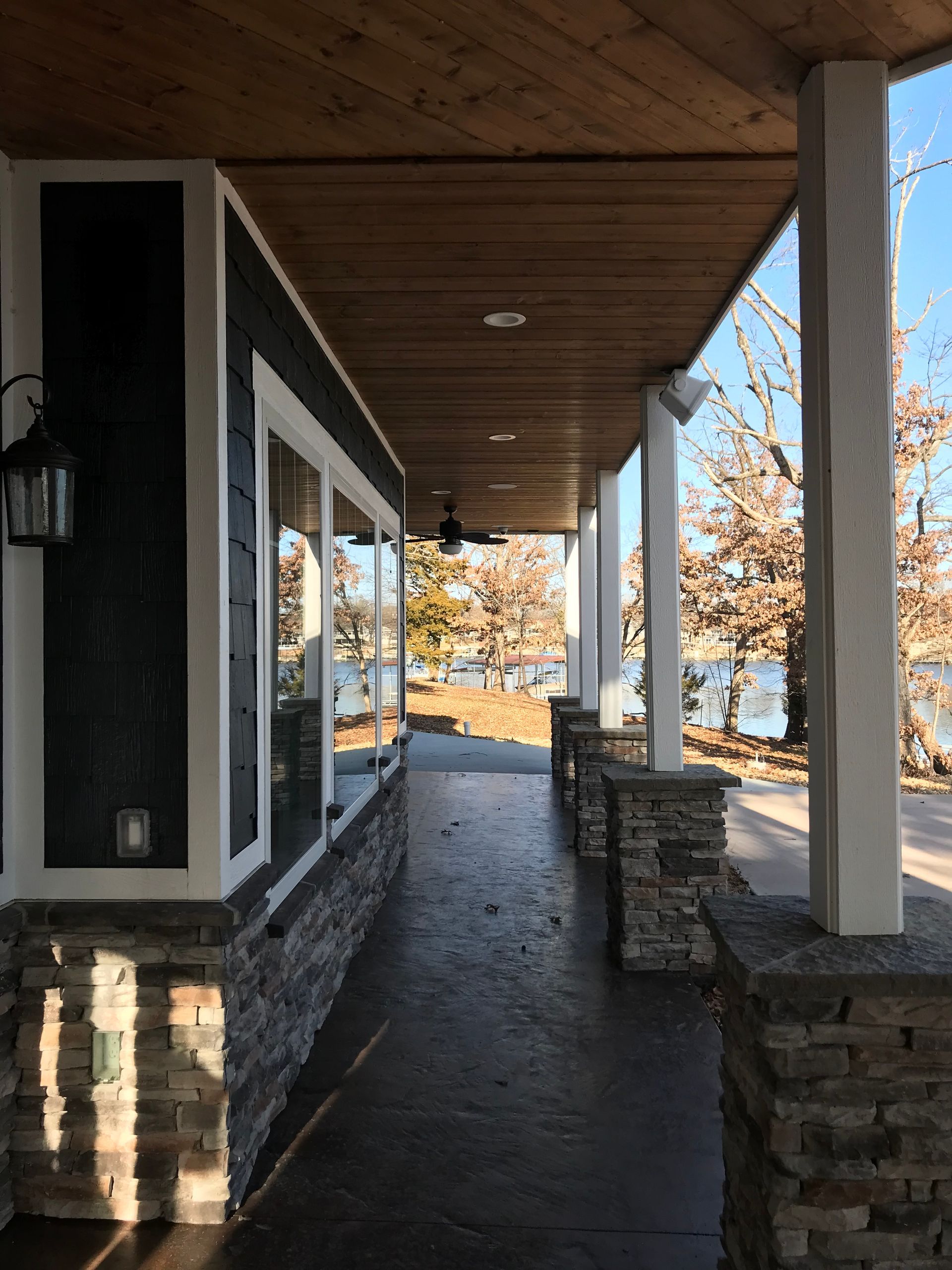 Covered front porch with stone columns, dark siding, and a walkway leading to autumn trees.
