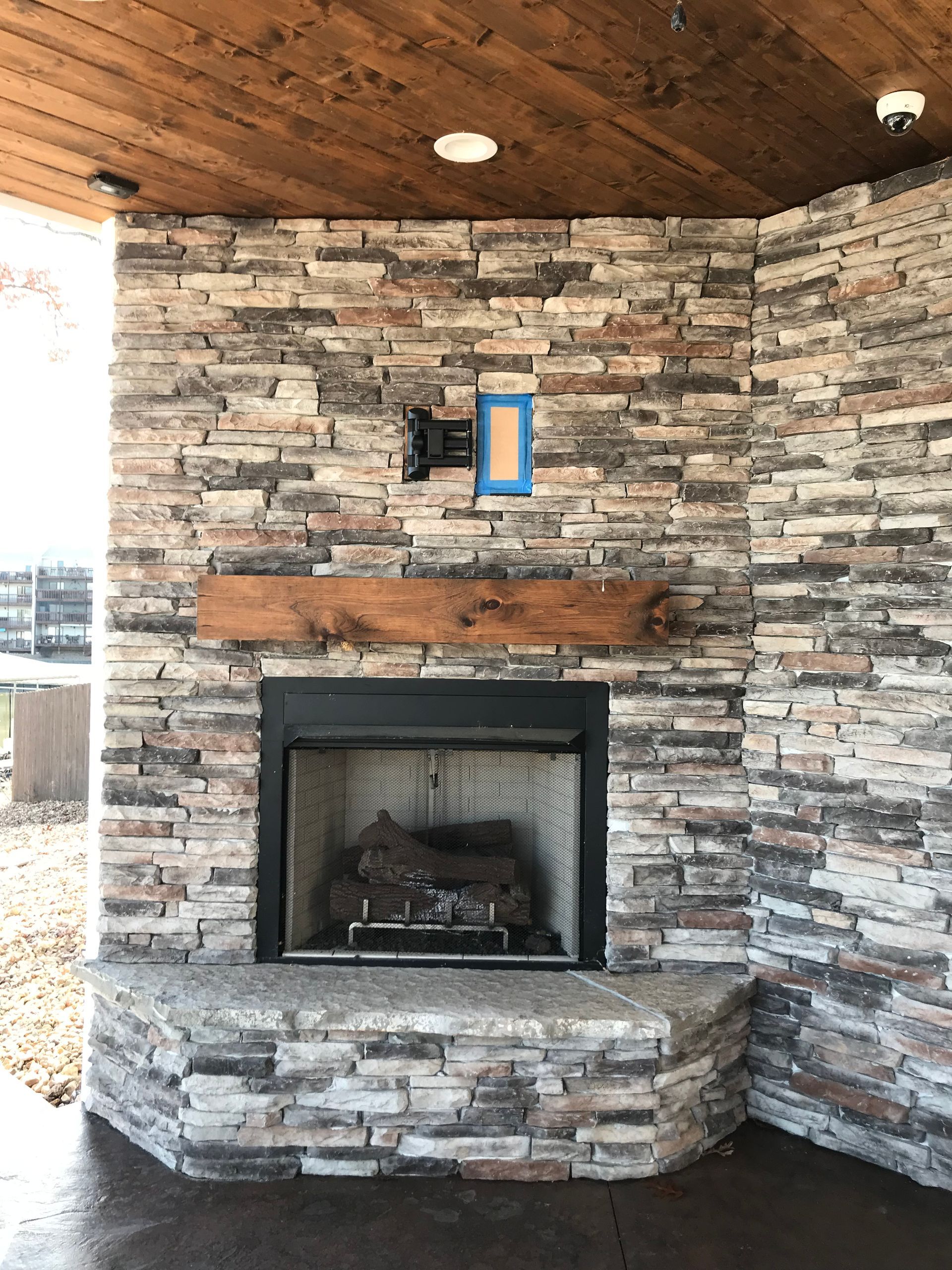 Stone fireplace with wooden mantel under a wood-paneled ceiling on a covered patio.