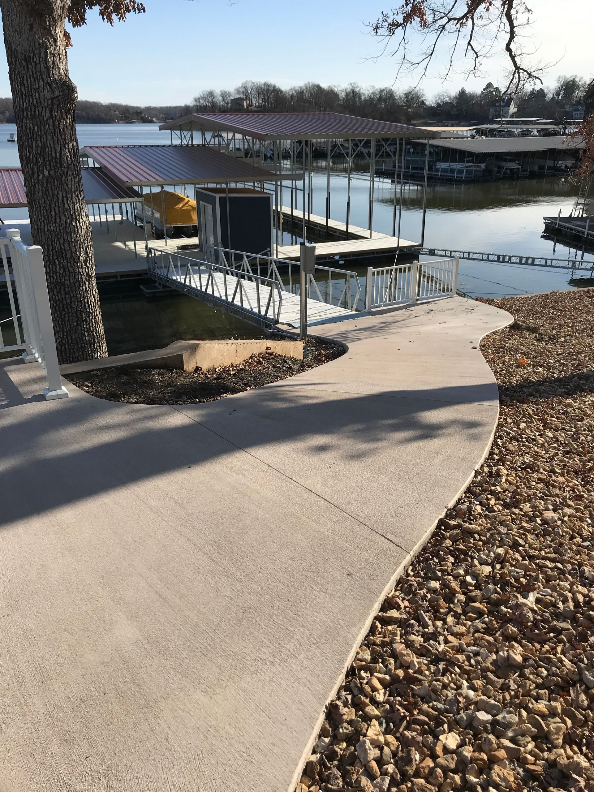 Curved walkway leading to a lakeside dock with a metal-roofed shelter and boats in the distance.