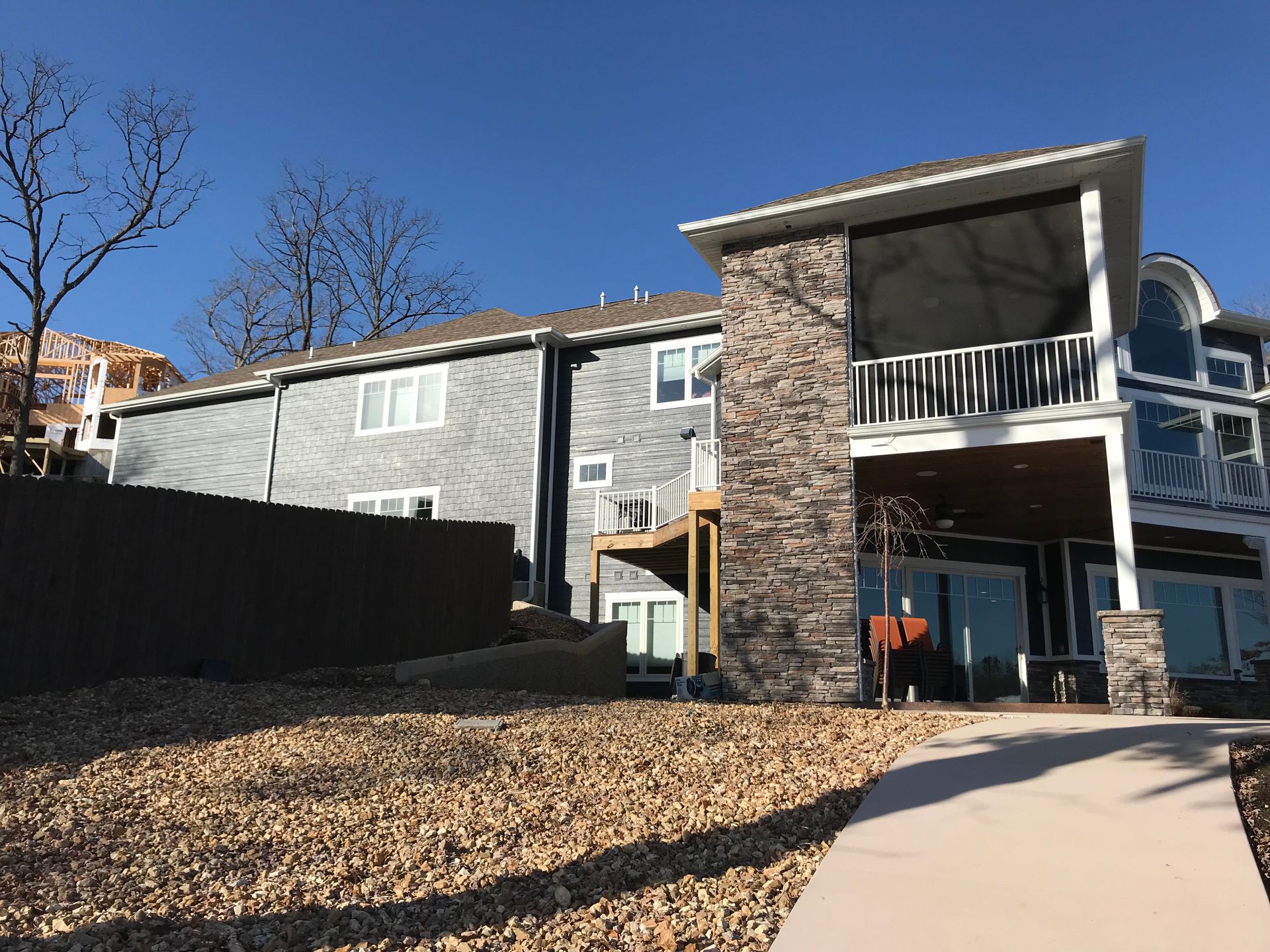 Apartment building with stone columns and balconies beside a gravel yard and paved walkway on a clear day