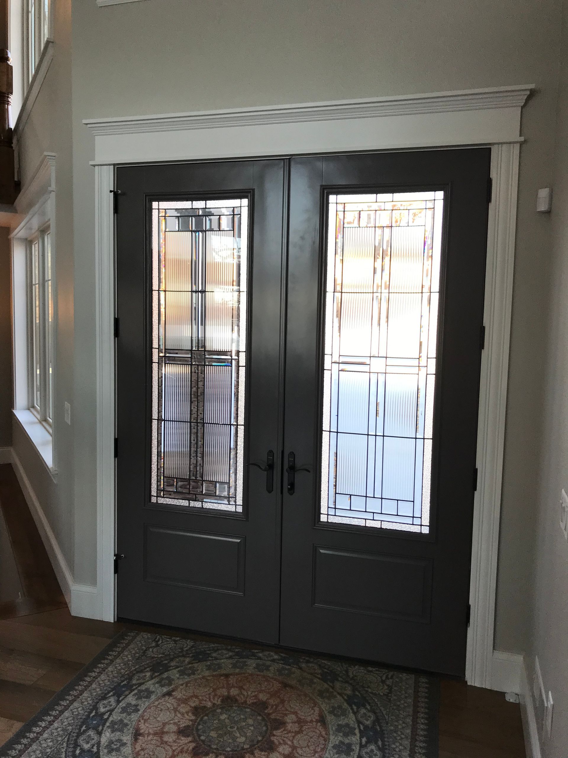 Dark double front doors with glass panels in a home entryway, framed by white trim and a patterned rug.