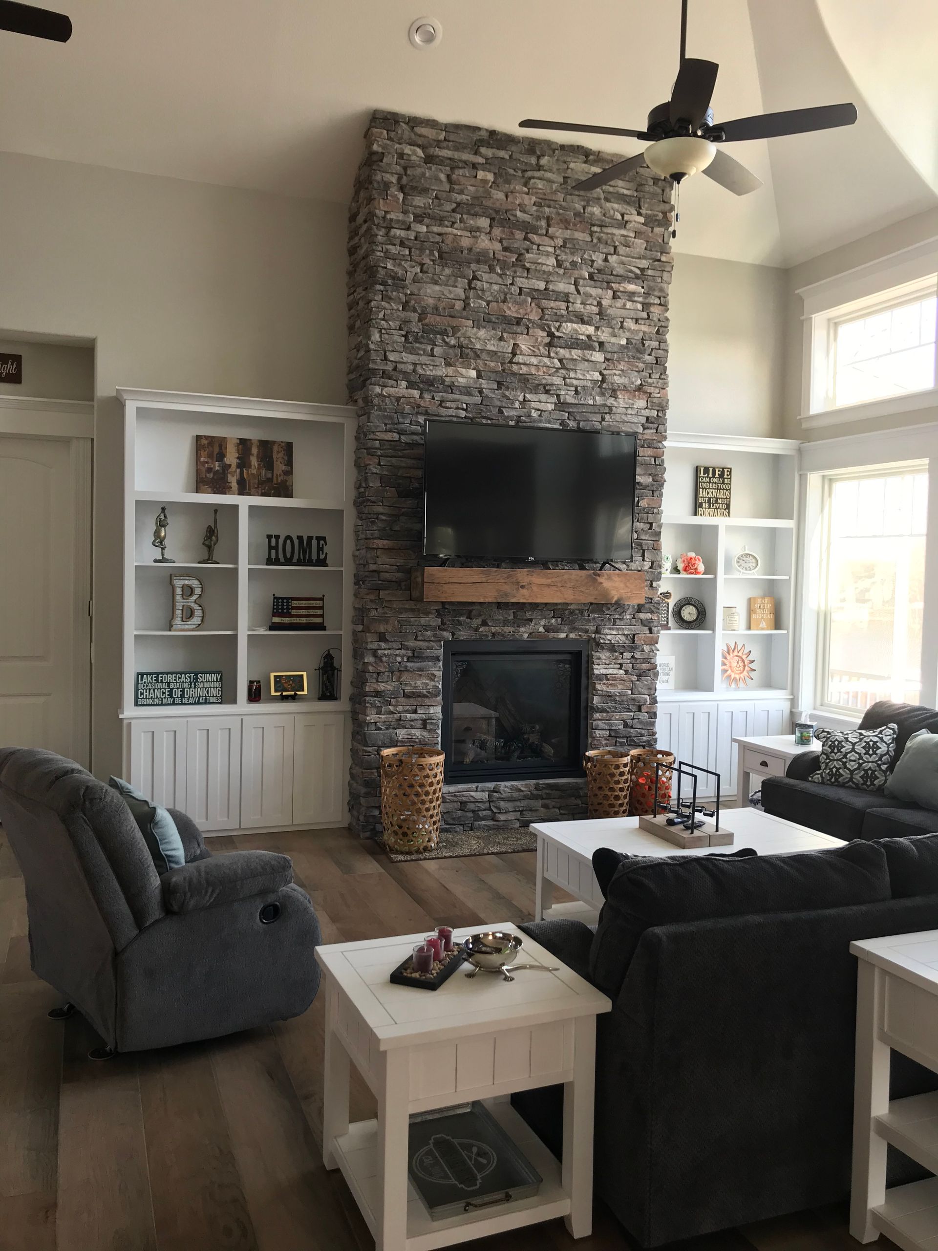 Living room with stone fireplace, TV, built-in shelves, gray seating, and large windows