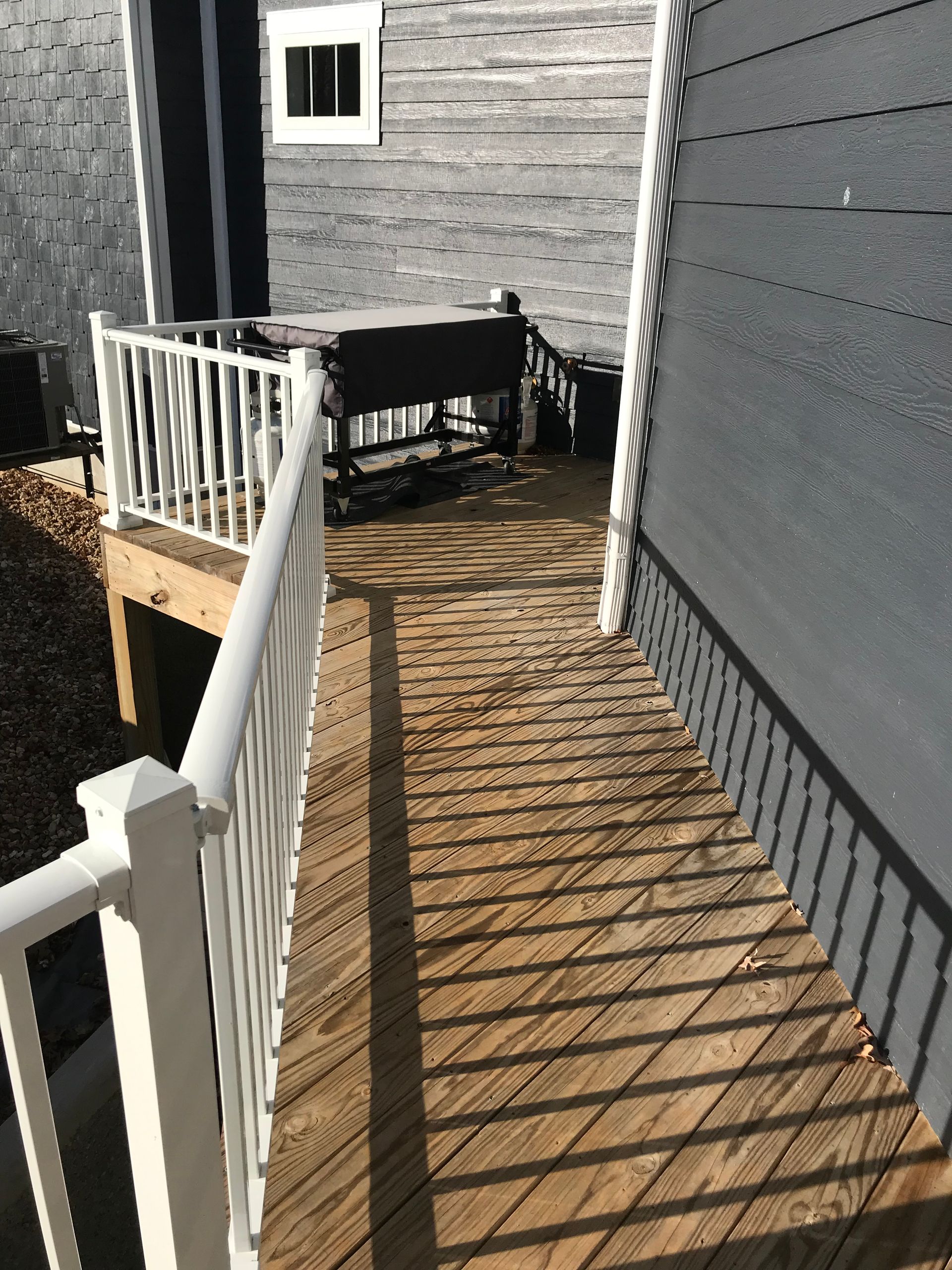 Narrow wooden deck walkway beside gray house, with white railing and outdoor chair in sunlight.