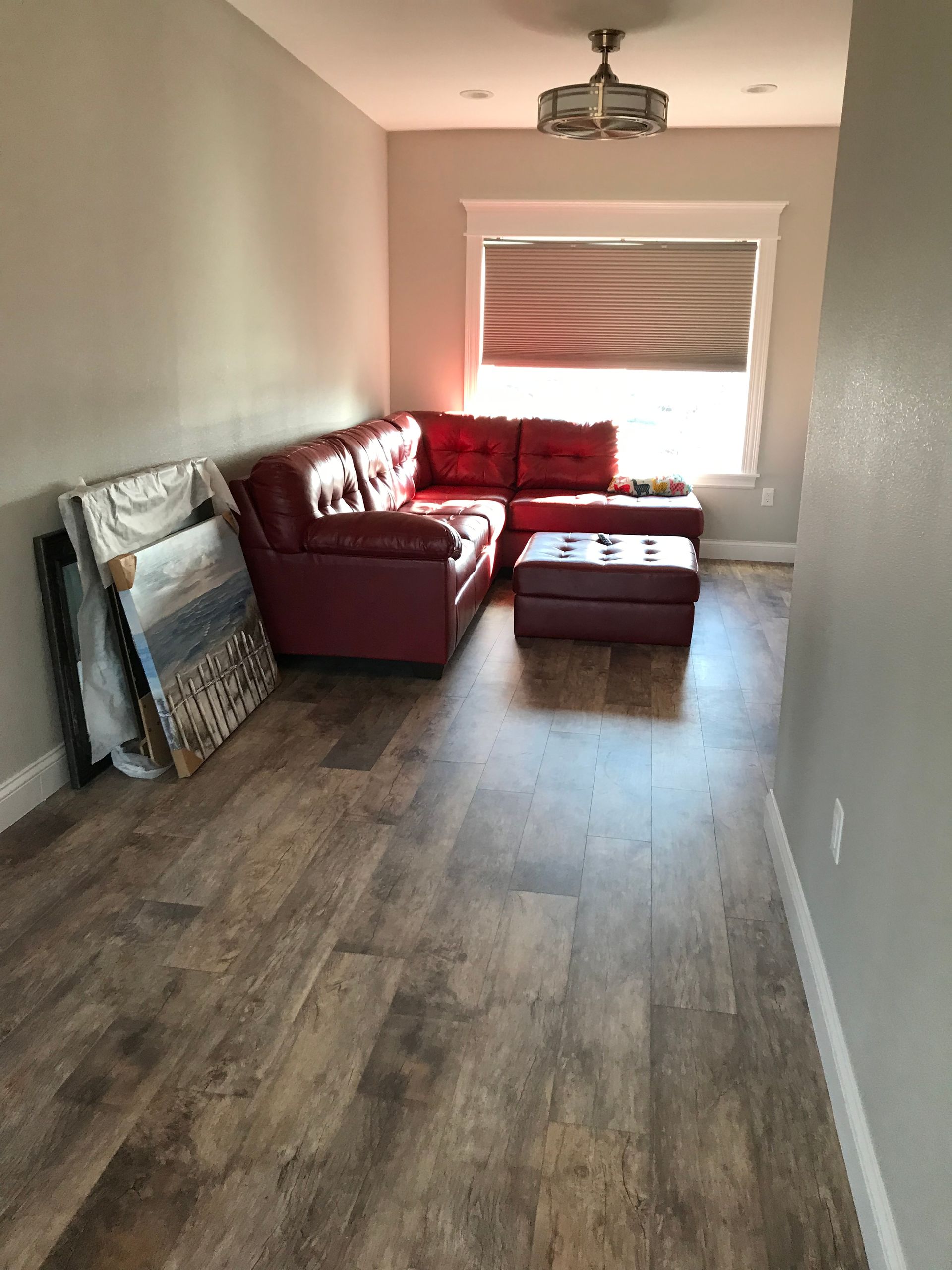 Empty living room with wood floors, red sectional sofa, ottoman, and large window with blinds.