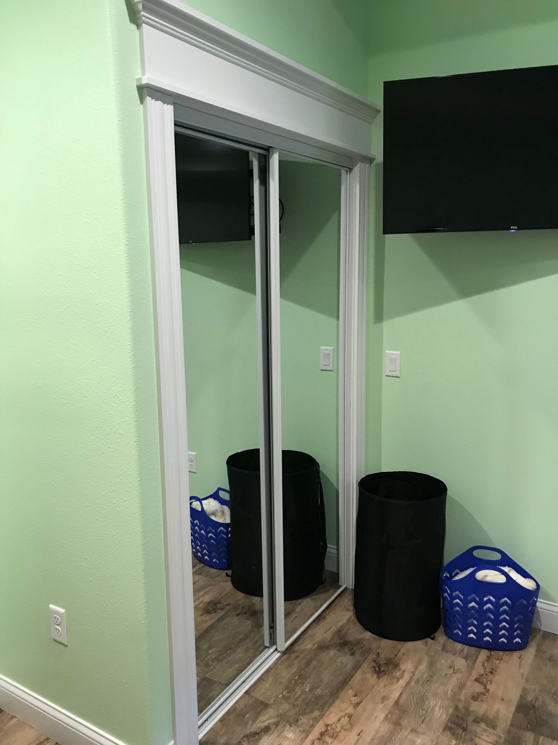 Green room with mirrored closet doors, wood floor, and a wall-mounted TV above laundry baskets and trash bins.