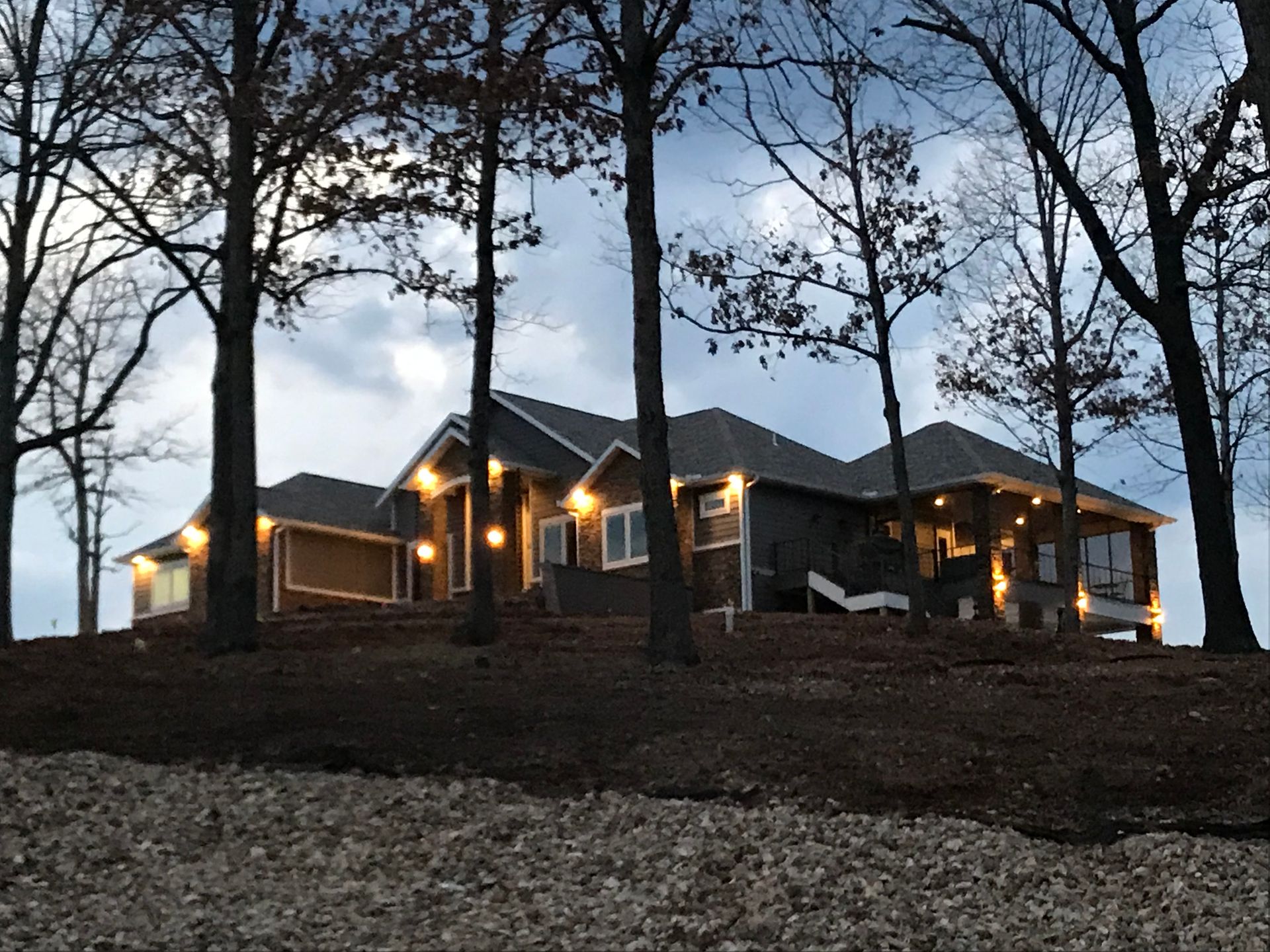 Lit house on a wooded hillside at dusk, seen through silhouetted trees.