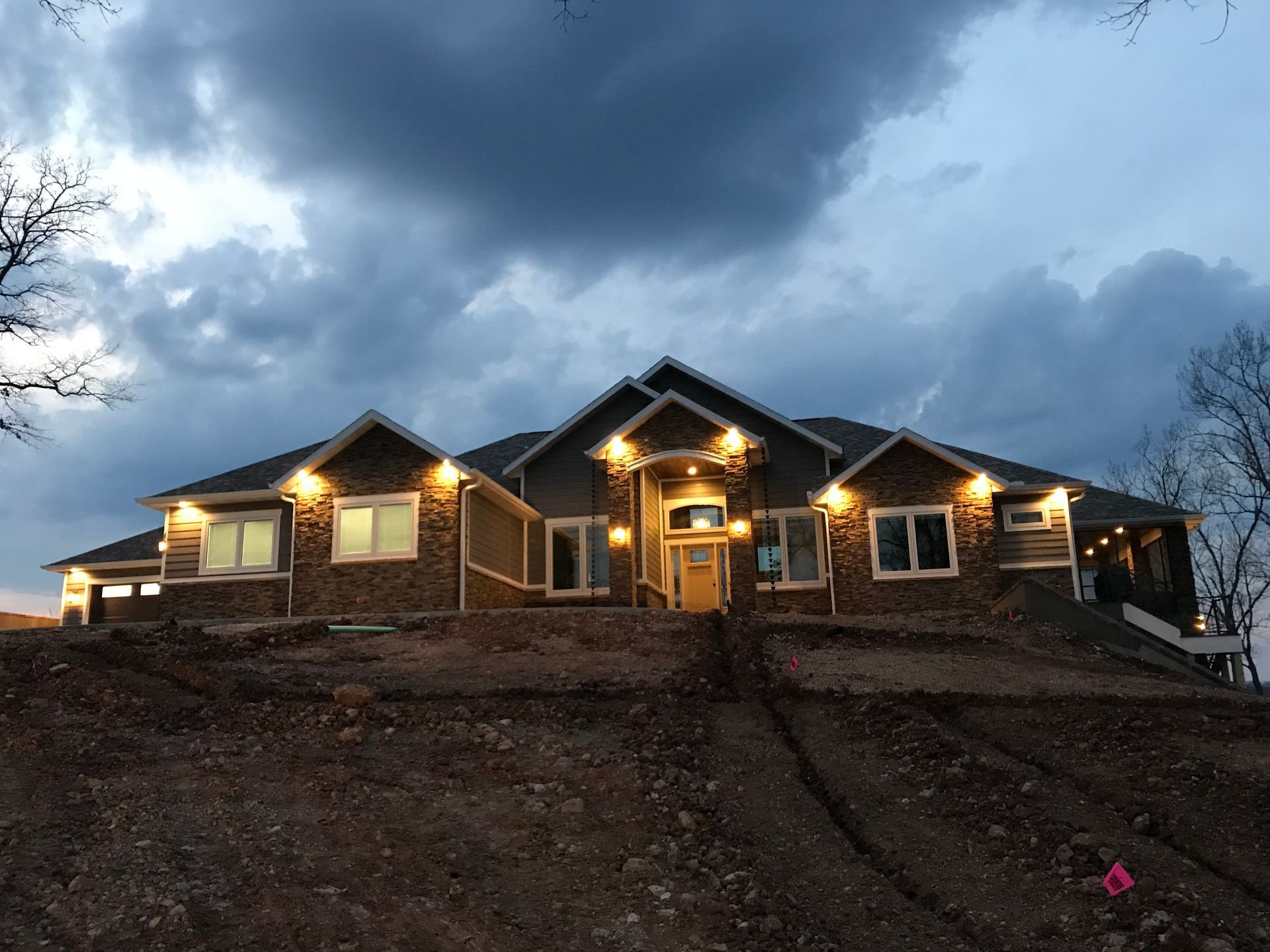 Two-story house glowing at dusk under dark storm clouds, on a hillside with bare trees.