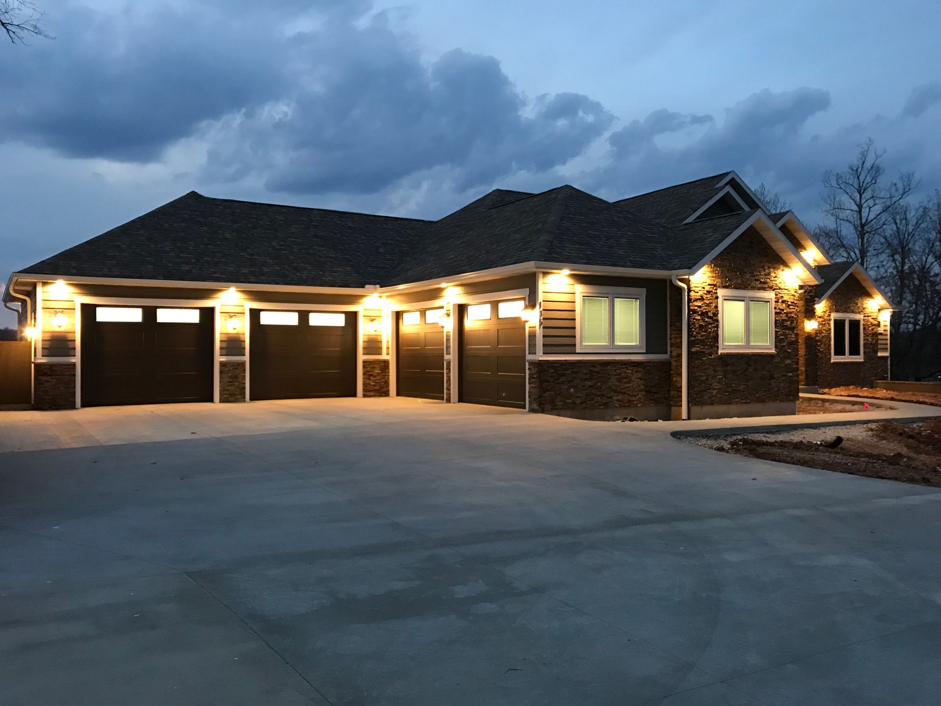 Large lit suburban house with three garage doors at dusk under a cloudy sky