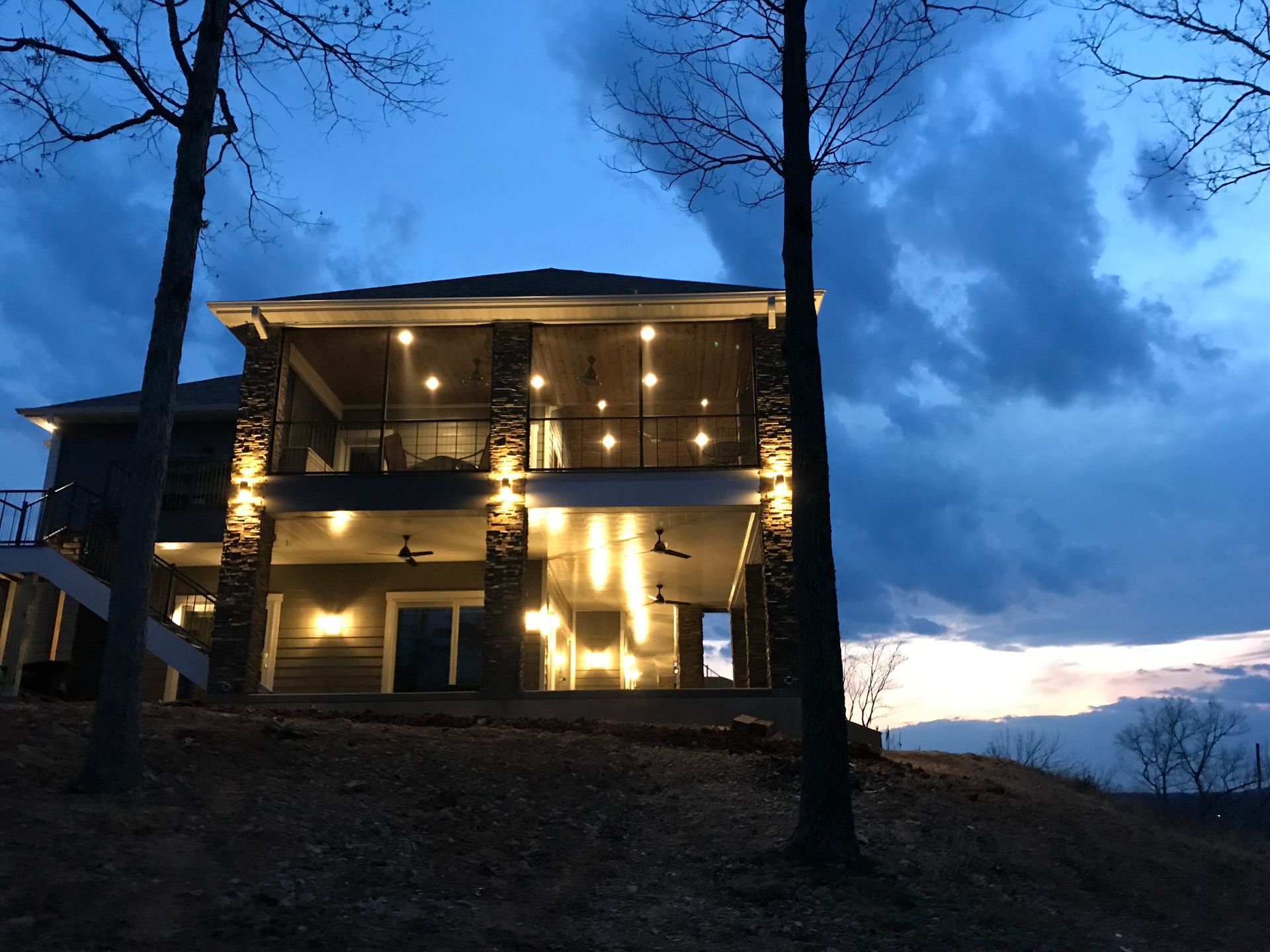 Illuminated two-story house at dusk with warm lights, trees in foreground, and cloudy blue sky.