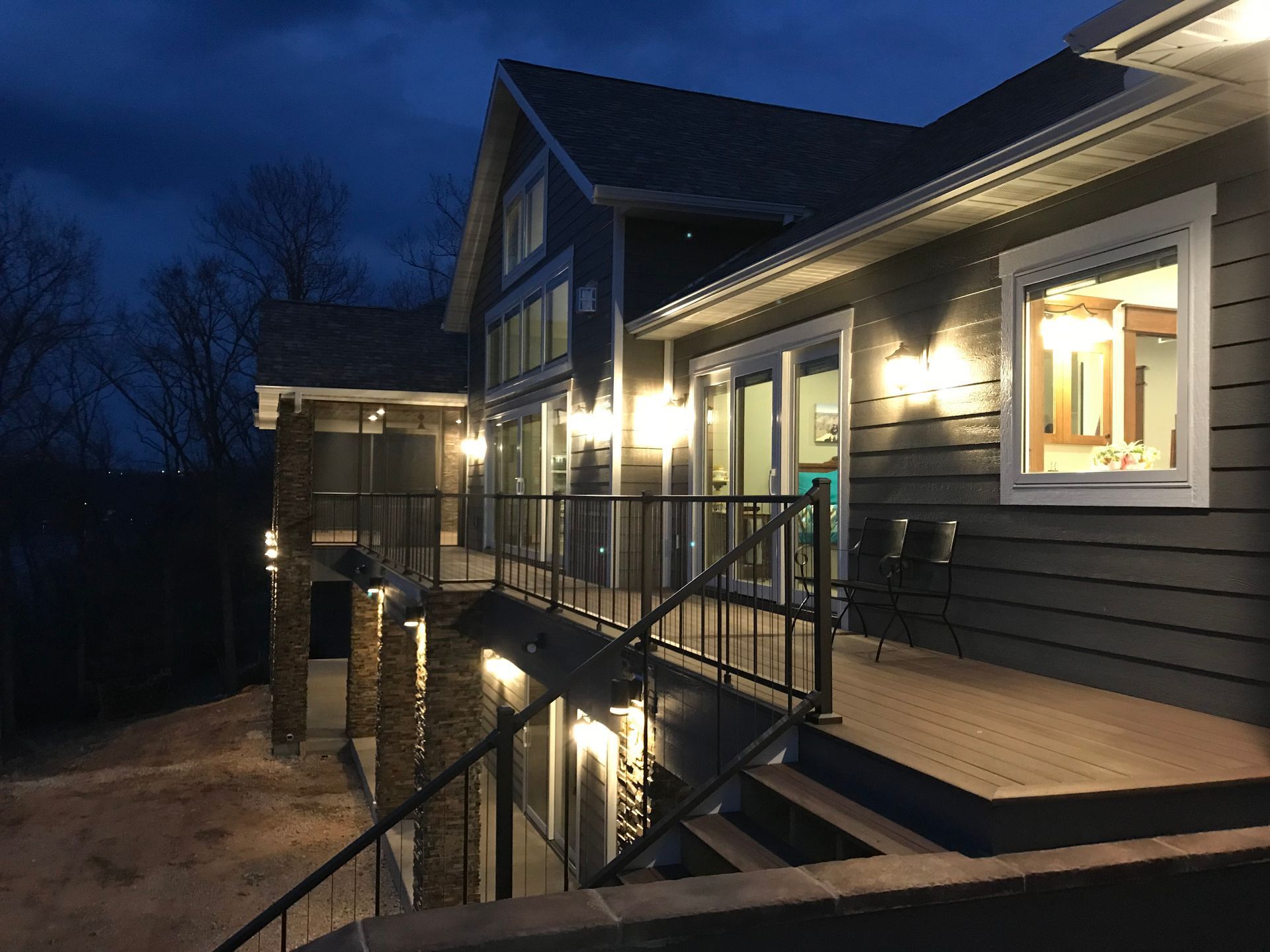 Nighttime view of a modern house deck with lit windows, railings, and stairs on a snowy slope