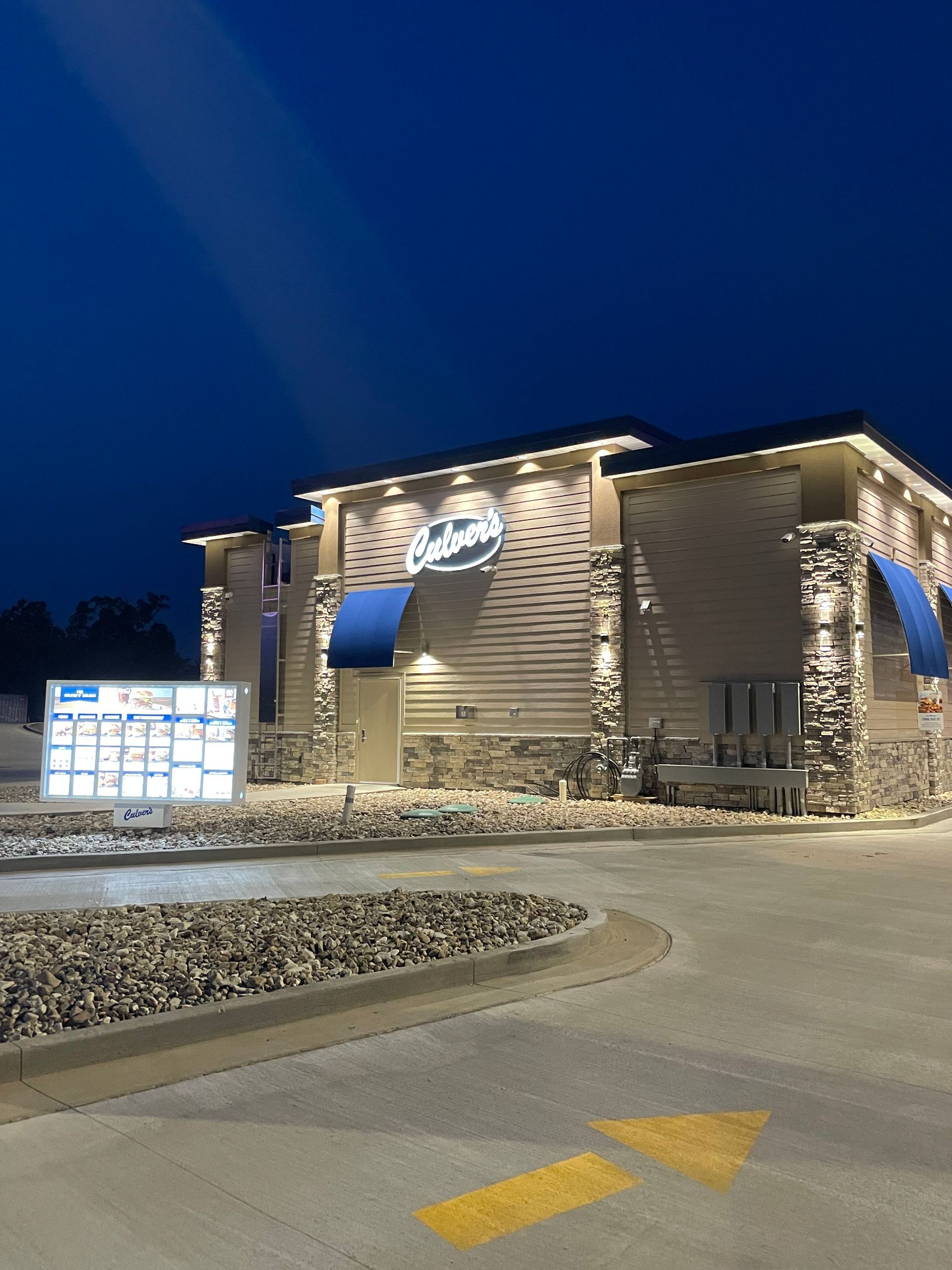 Nighttime view of a lit stone building with a blue awning, signboard, and curved parking lot entrance.
