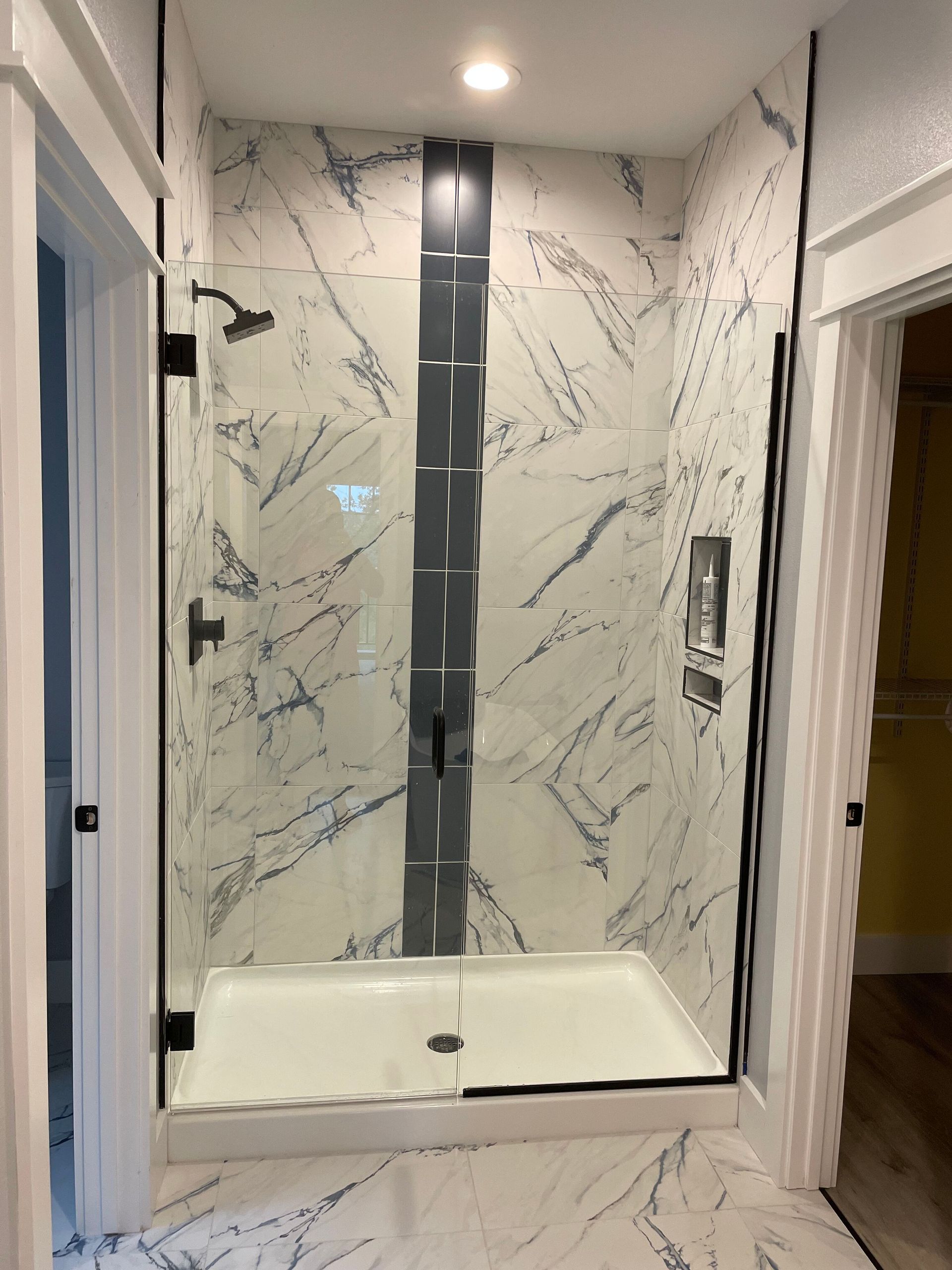 Small marble-tiled shower with glass door, white base, and black fixtures in a narrow bathroom corridor