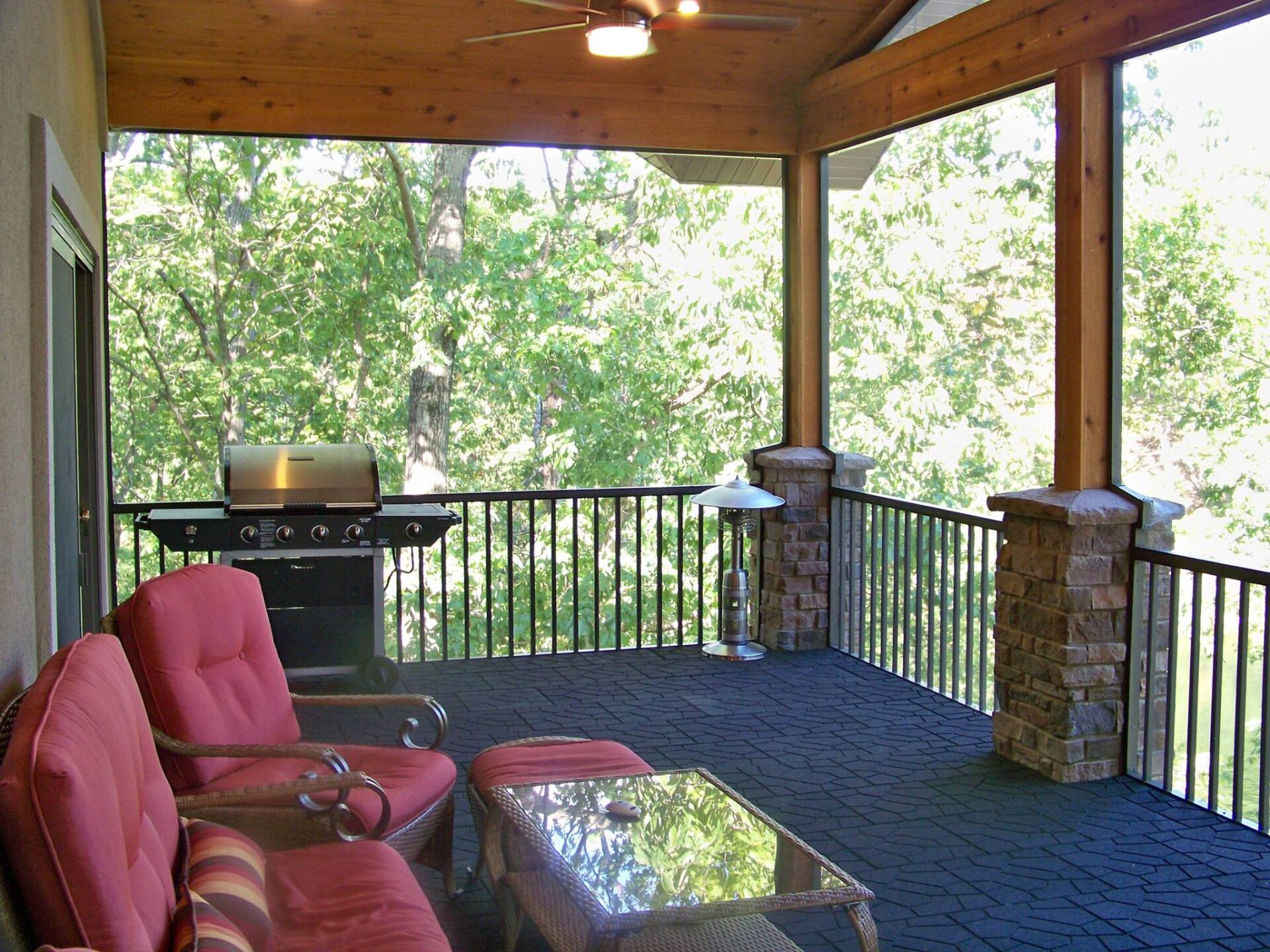 Covered porch with red cushioned chairs, glass table, and grill overlooking trees and black railing.
