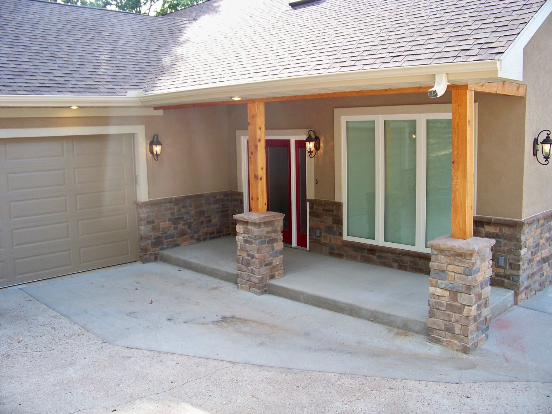 Front entry of a beige house with stone columns, garage door, and large sliding glass doors under a shingled roof