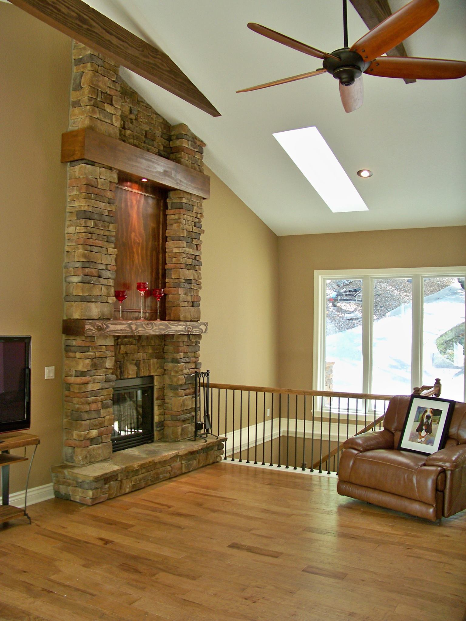 Living room with stone fireplace, hardwood floors, ceiling fan, and brown leather chair by a bright window.