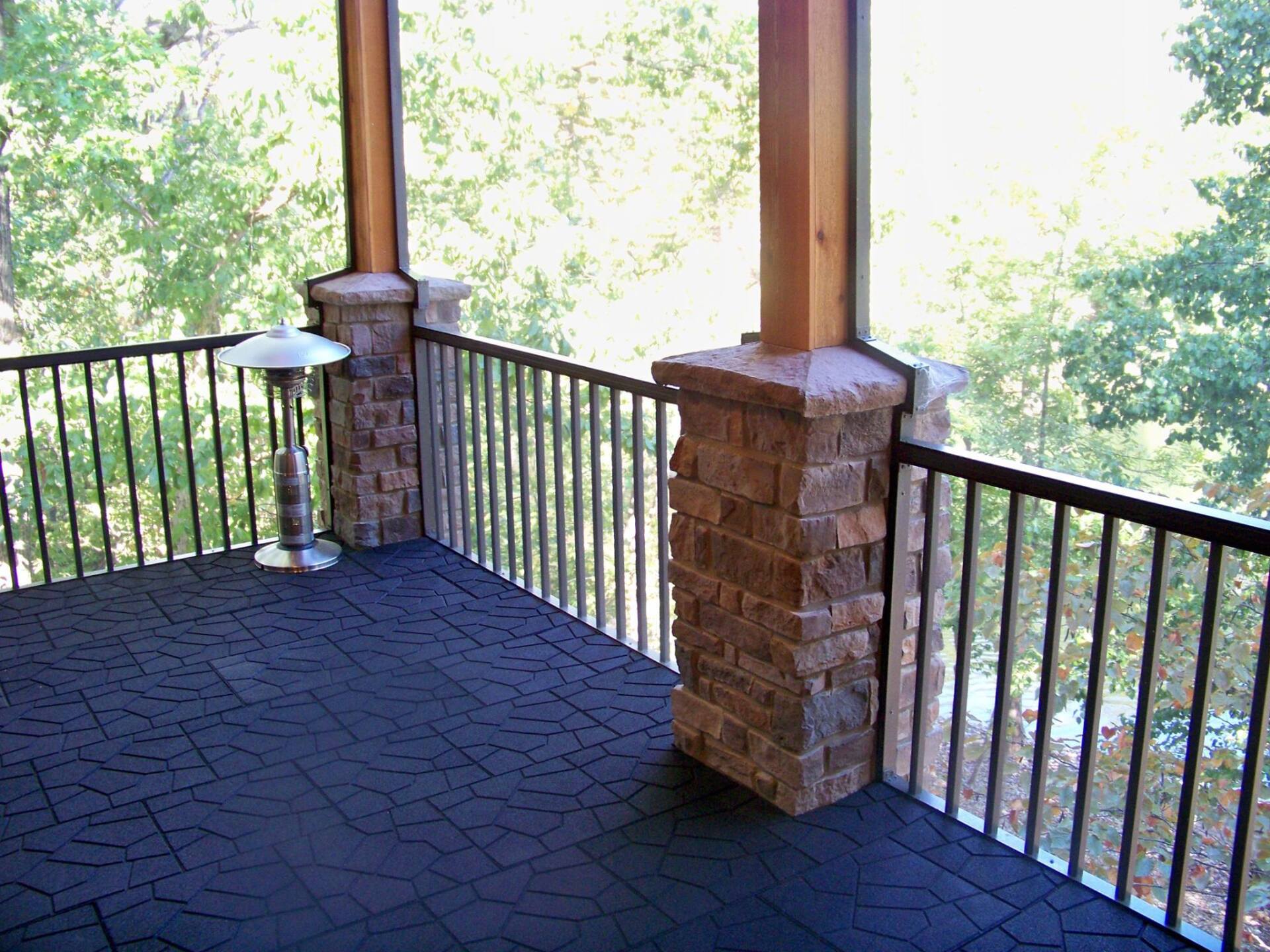 Covered porch with black railings, stone columns, and a dark patterned floor overlooking trees.
