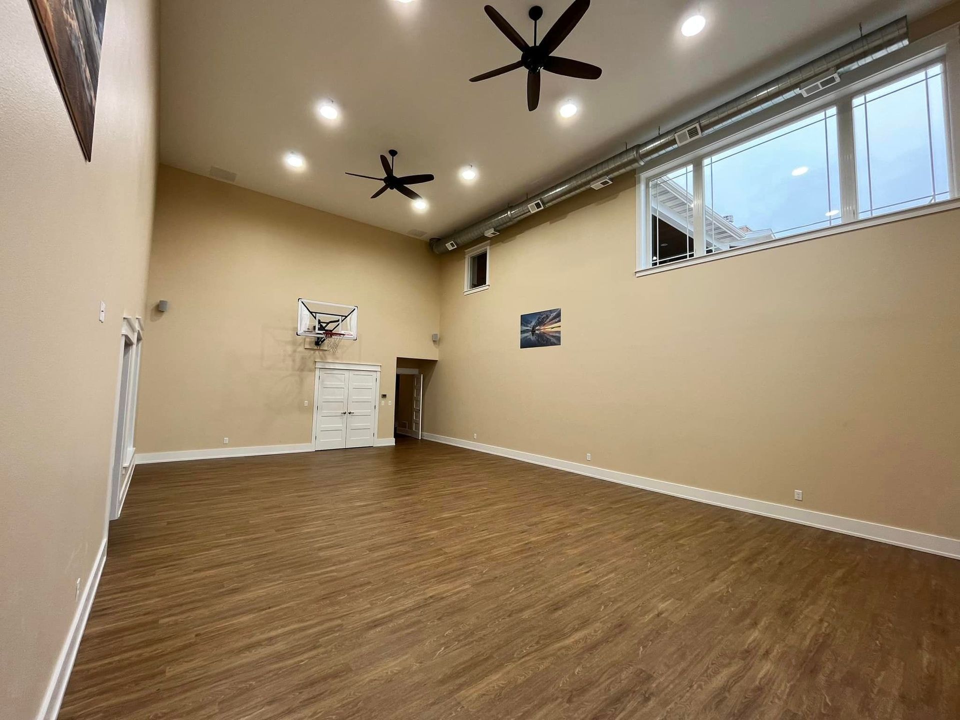 Empty beige room with wood-look floor, recessed lights, ceiling fans, and a small high window.
