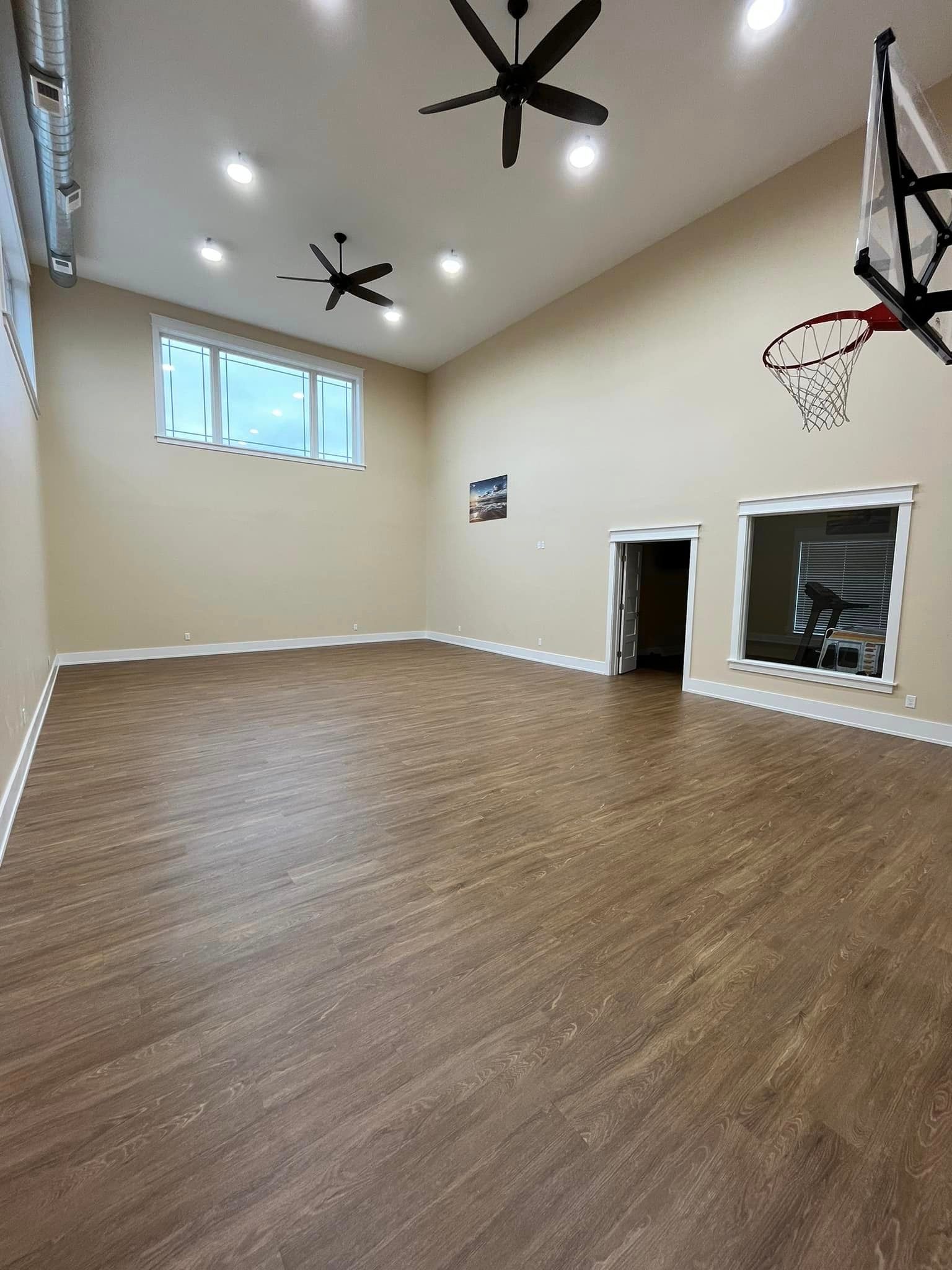Empty indoor basketball court with wood floor, high ceilings, ceiling fans, windows, and wall-mounted hoop
