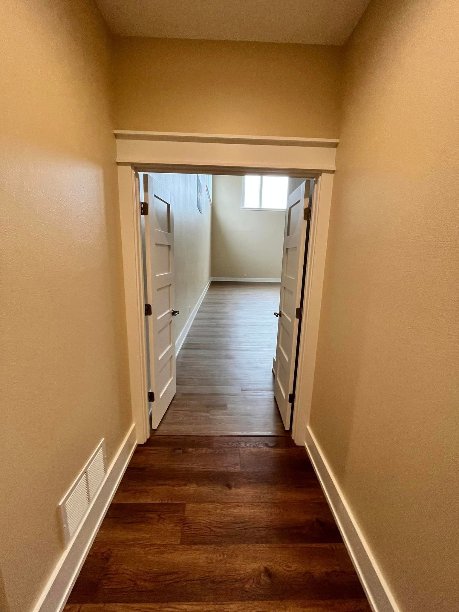 Narrow hallway with tan walls, dark wood floor, and open white double doors leading to a bright room.