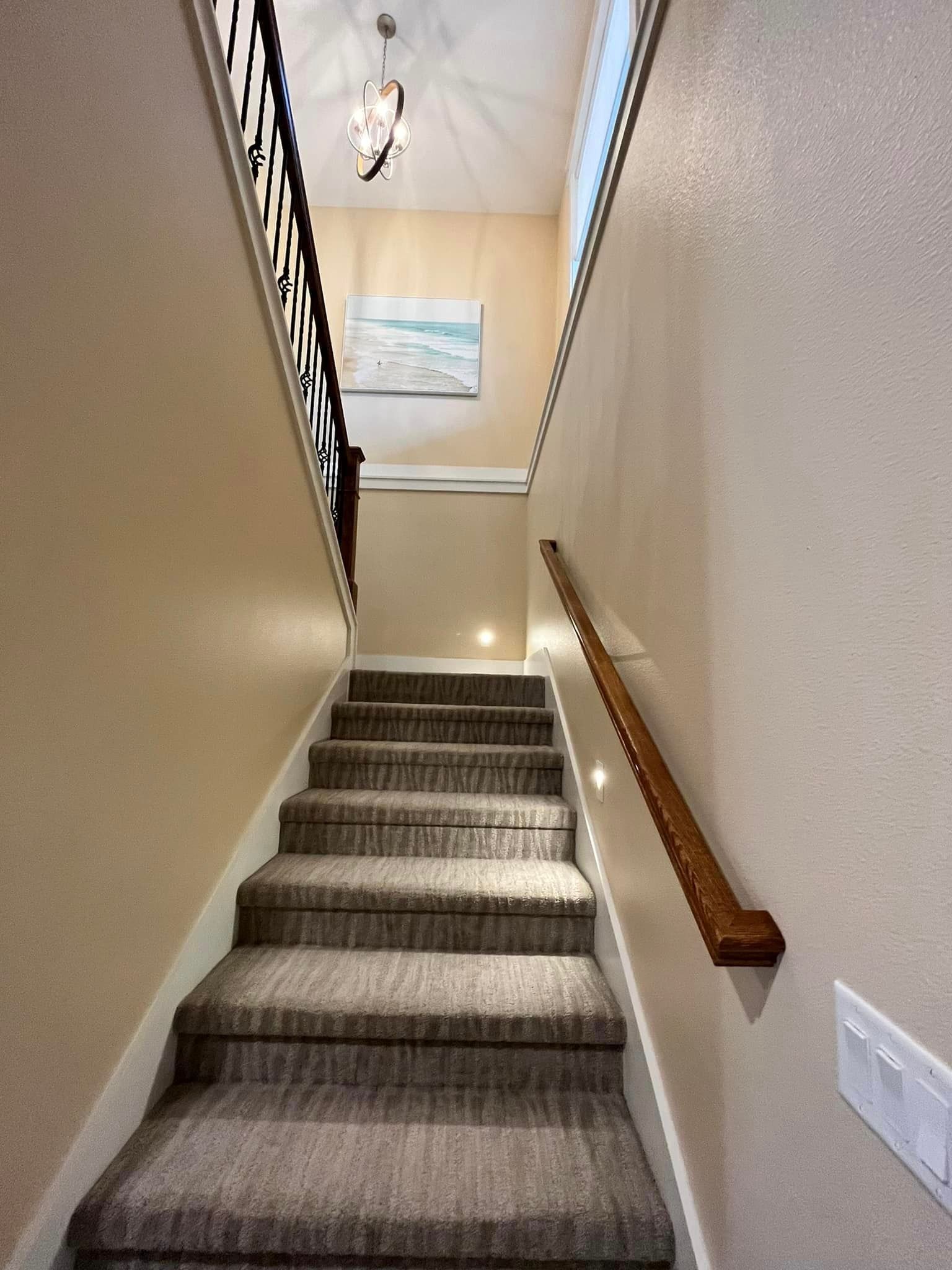 Carpeted indoor staircase with wooden handrail leading up to a landing with a hanging light and wall art.