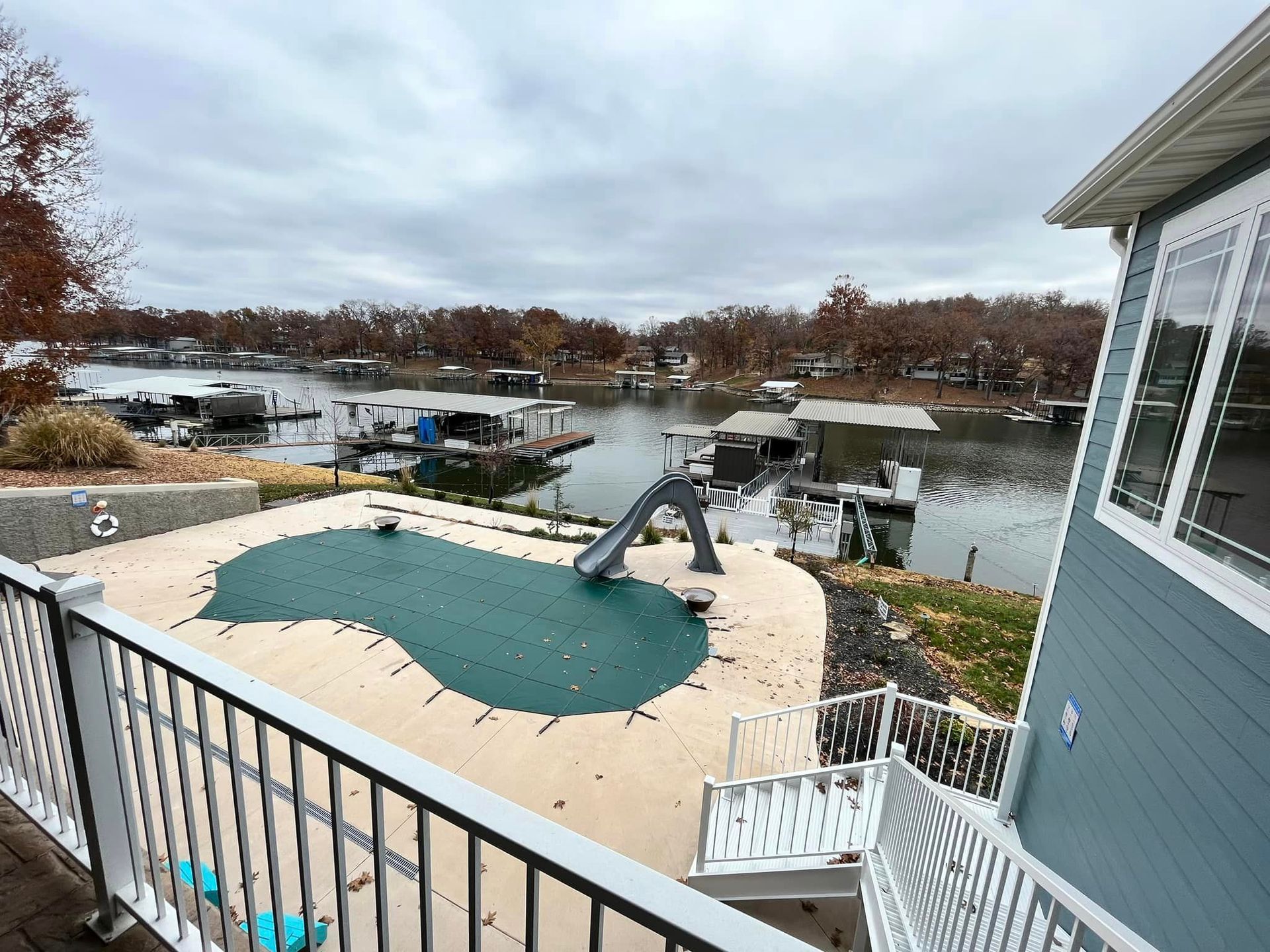 Backyard deck with an in-ground pool and slide overlooking a calm waterfront under cloudy skies