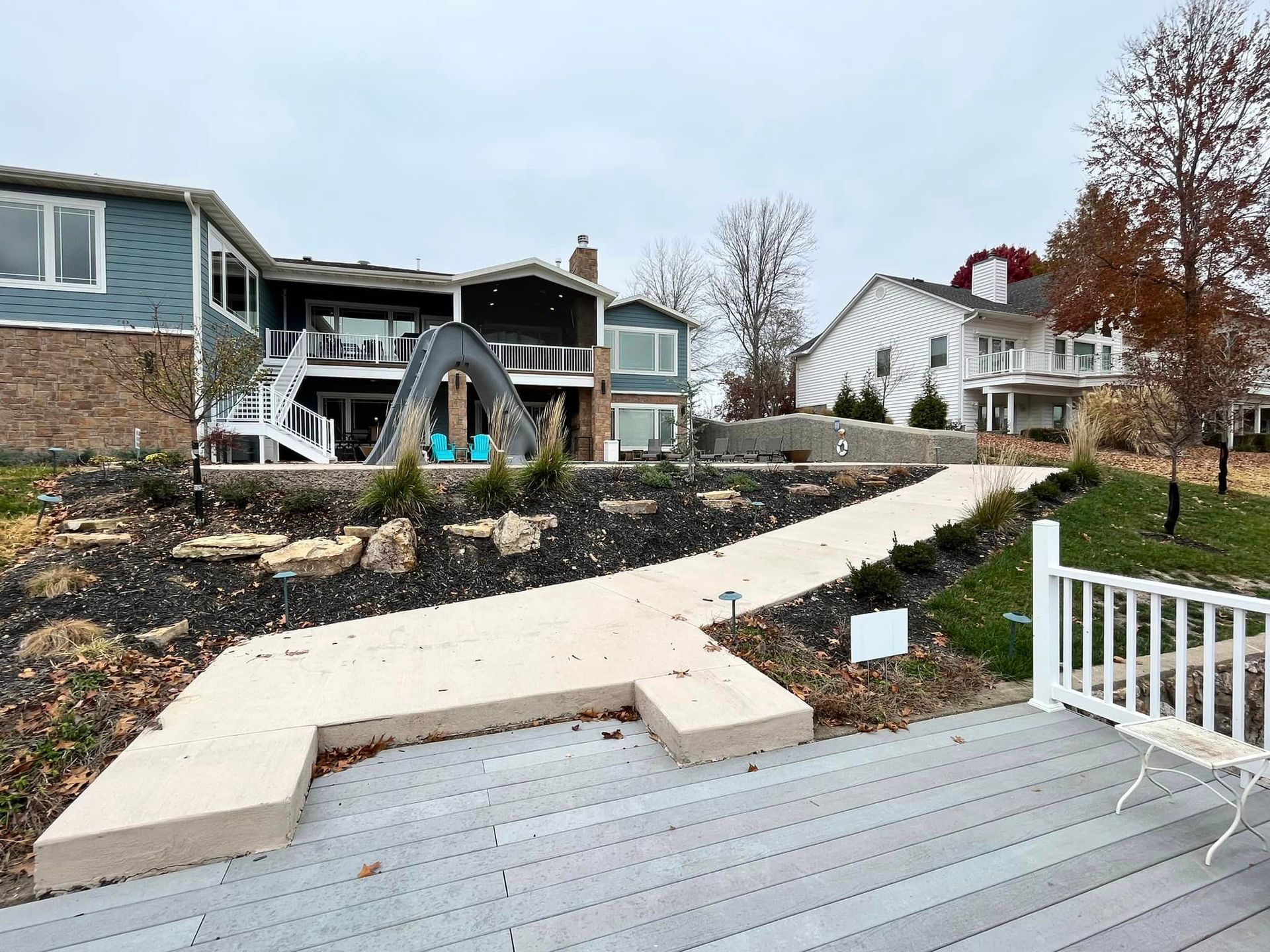 Backyard with a curved concrete path, deck, white fence, and houses under overcast sky