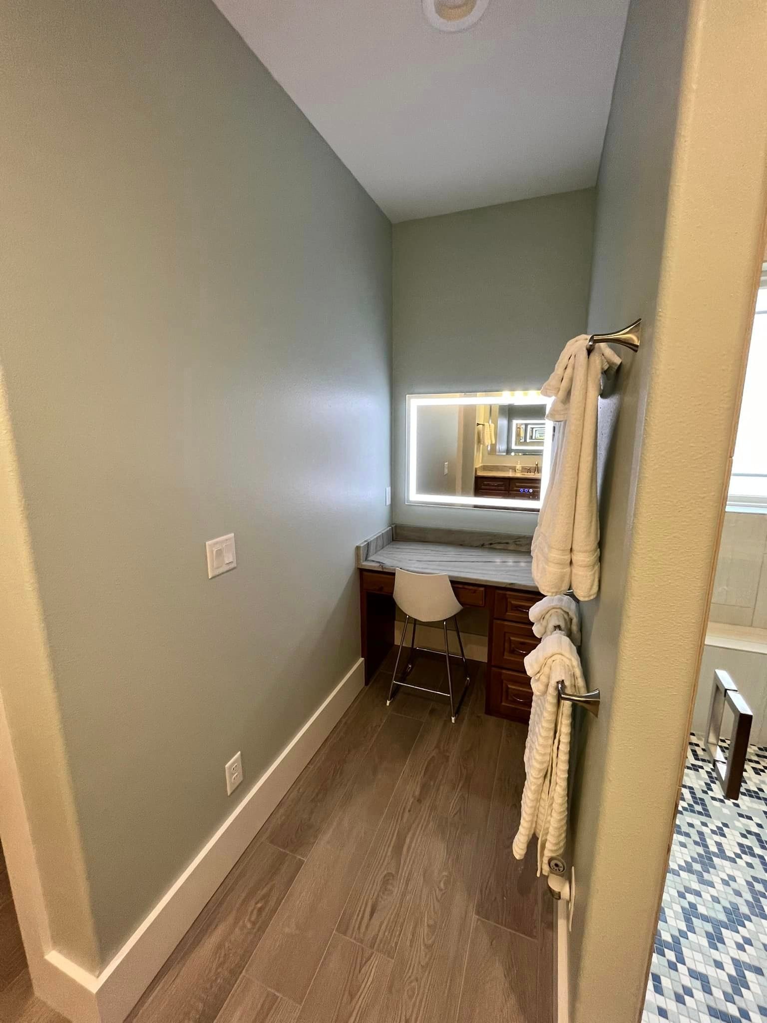 Narrow hallway with a small vanity desk, stool, mirror, and hanging robe beside a bedroom.