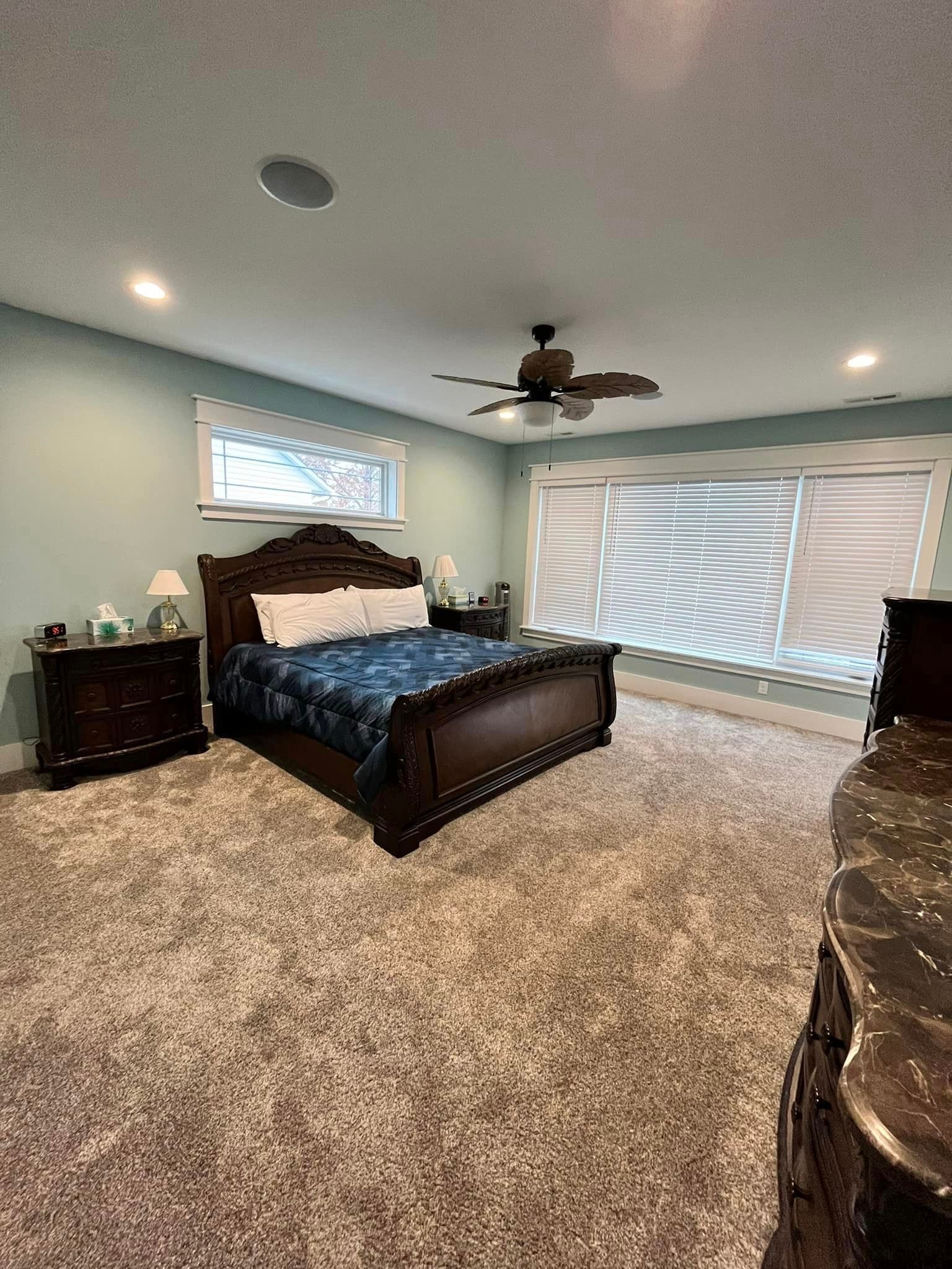Bright bedroom with dark wood bed, beige carpet, white blinds, and a ceiling fan.