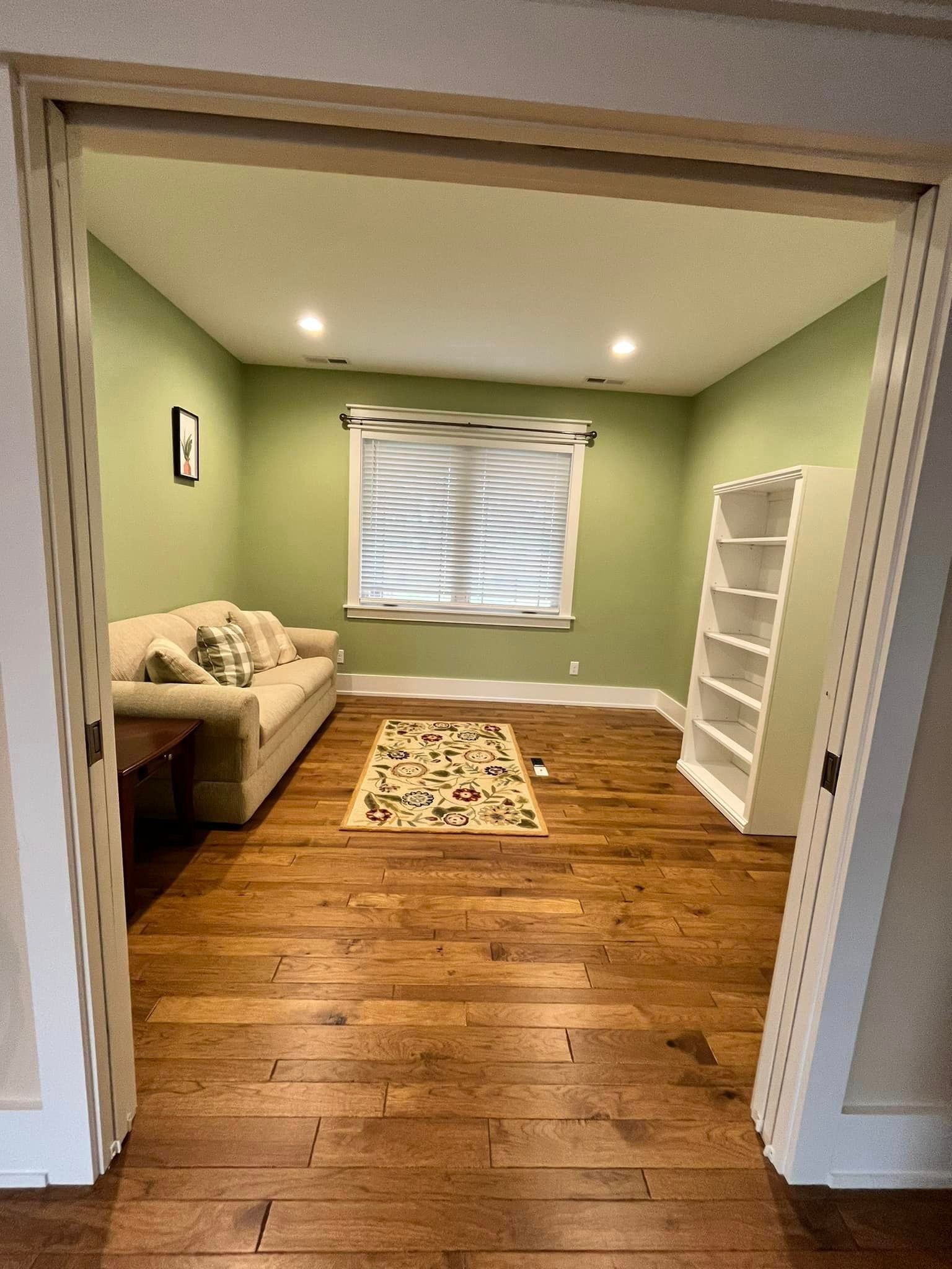 Small cozy room with green walls, wood floor, beige sofa, rug, window, and white shelving
