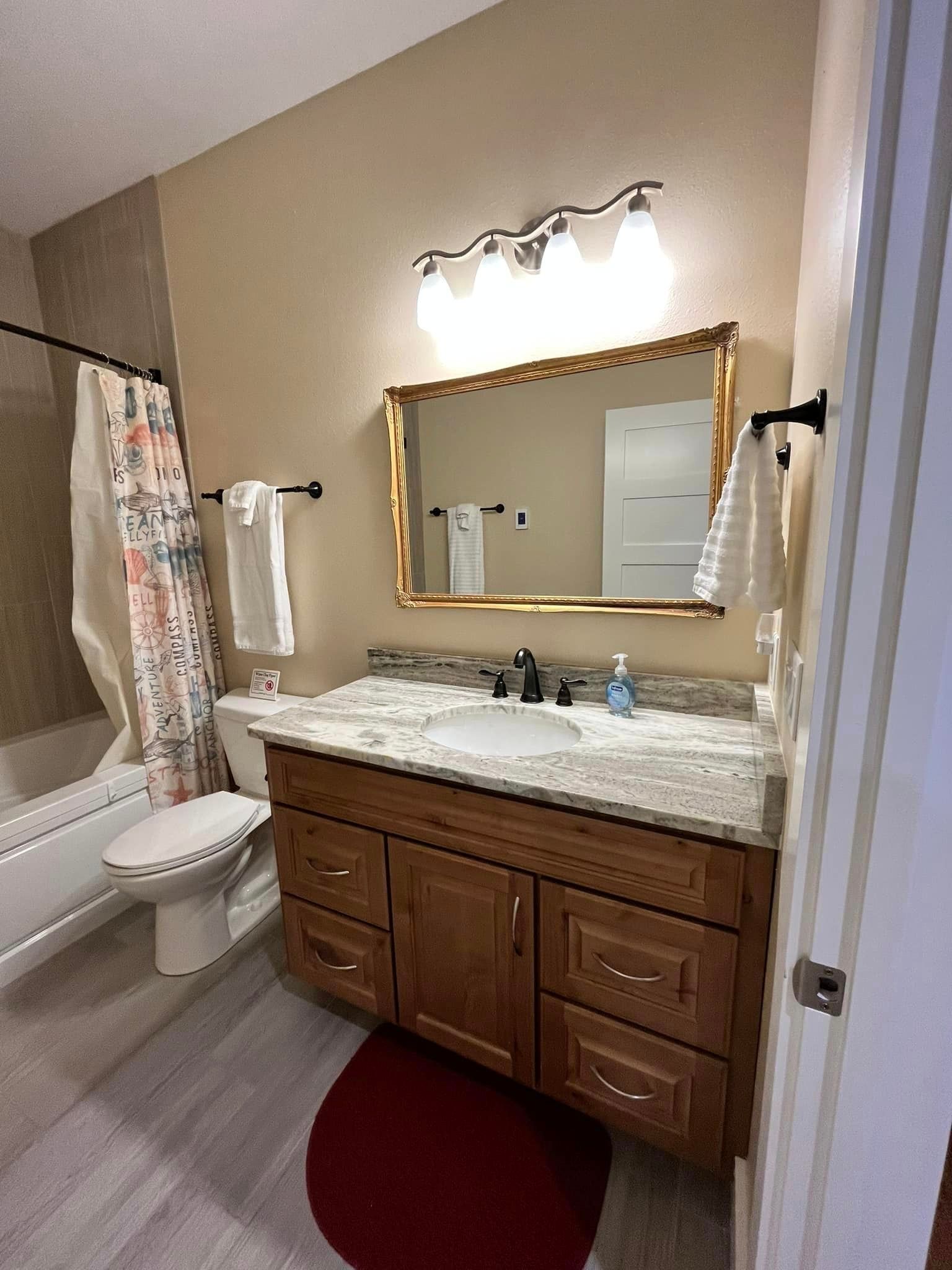 Small bathroom with a wood vanity, marble countertop, framed mirror, toilet, tub, and floral shower curtain.