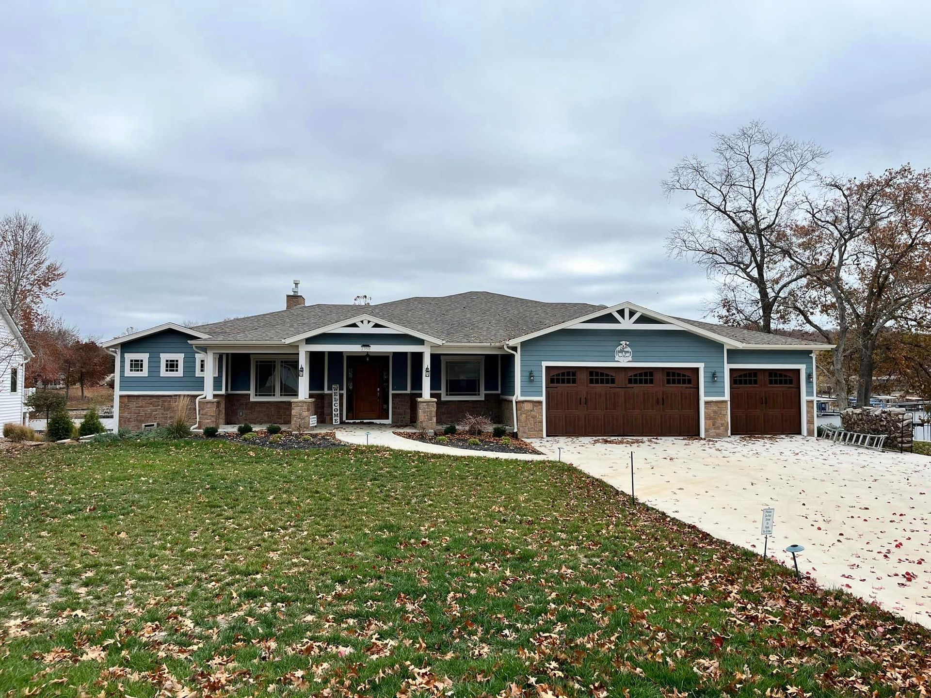 Suburban brick ranch house with a two-car garage, front porch, and a large leaf-covered lawn under cloudy skies