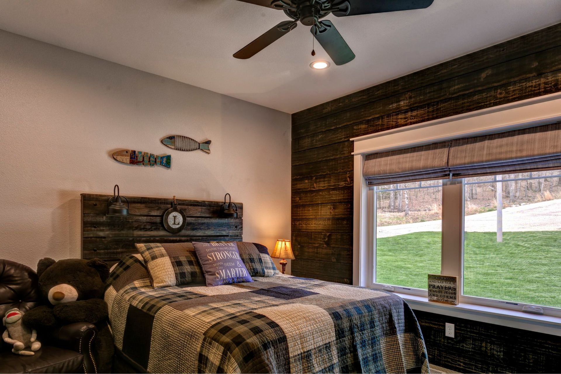 Rustic bedroom with plaid bedspread, teddy bear, wood accents, and a large window overlooking a yard